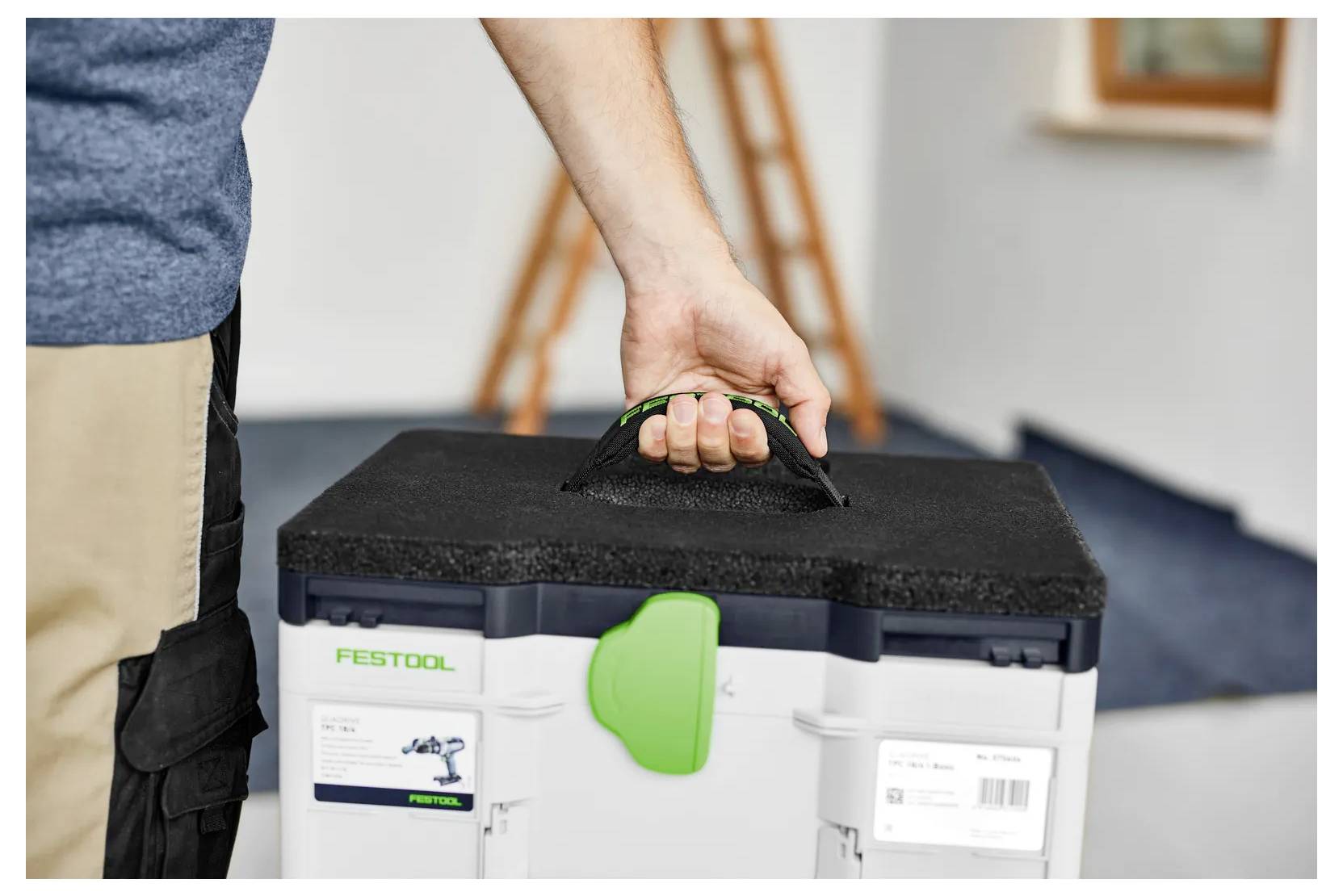 Person carrying a Festool tool case by its handle in a workshop or room interior.Eine Person trägt einen Festool-Werkzeugkoffer an seinem Griff in einer Werkstatt oder einem Innenraum.