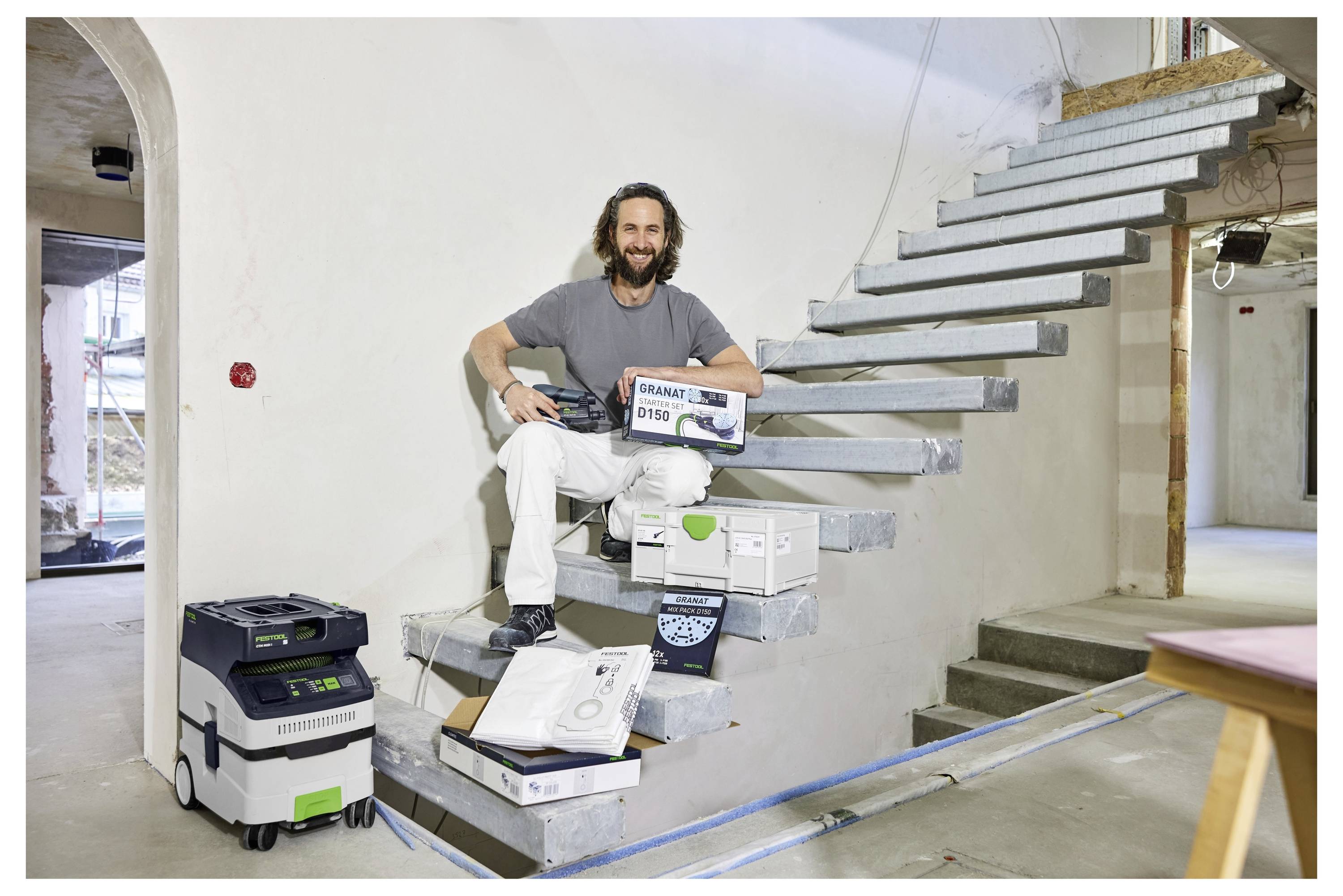 Ein Mann lächelt, während er auf unfertigen Betontreppen auf einer Baustelle sitzt, hält ein Elektrowerkzeug und einen verpackten Gegenstand, mit anderen Werkzeugen in der Nähe.
