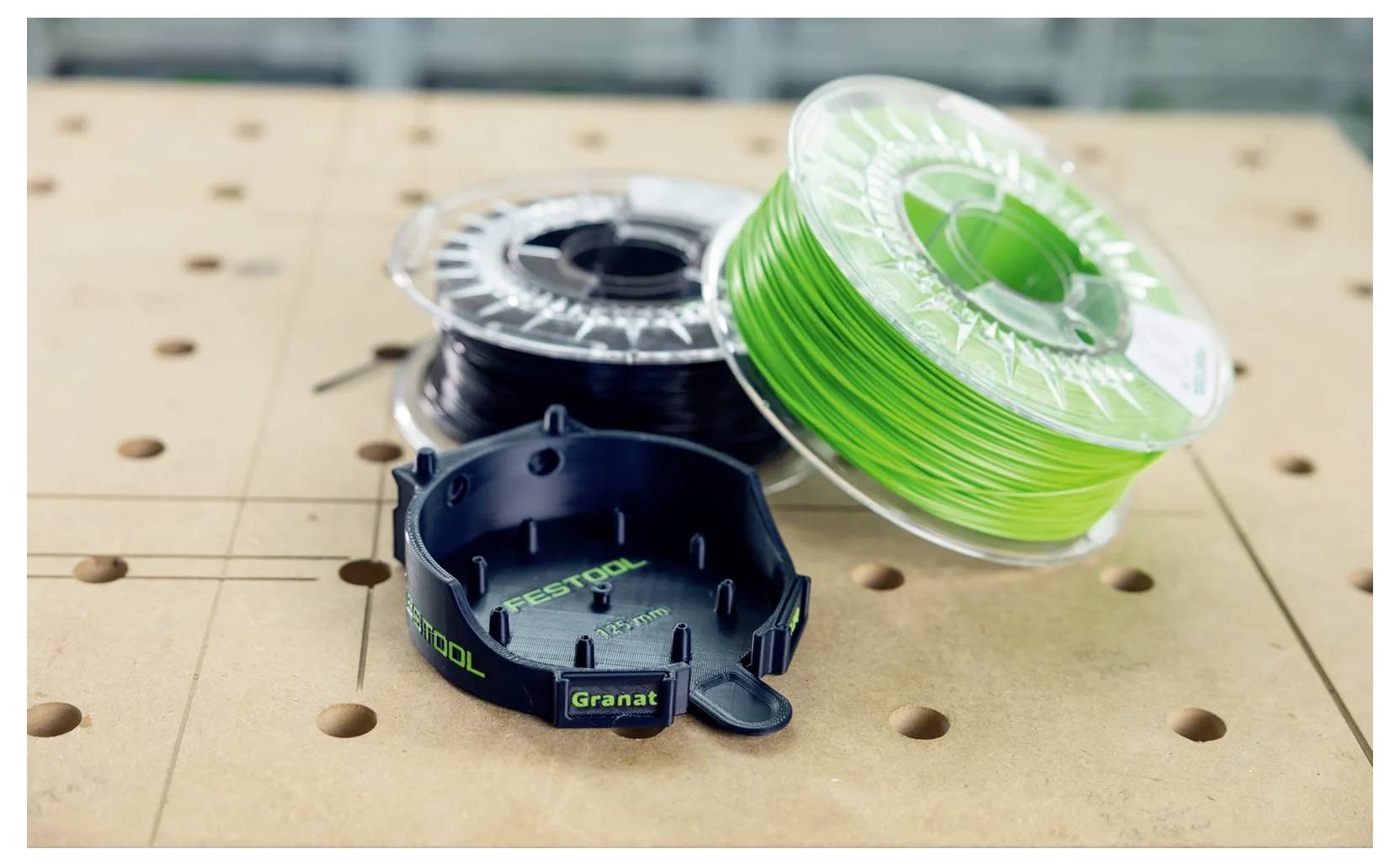 Drei Spulen mit 3D-Druckerfaden auf einer perforierten Werkbank, einschließlich schwarzem und hellem grünem Kunststofffaden.