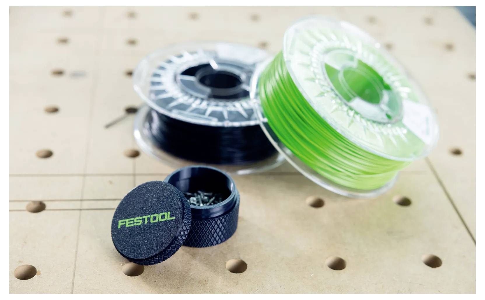 Drei Spulen durchsichtiges Filament und ein schwarzer Festool-Behälter stehen auf einer perforierten Werkbank.