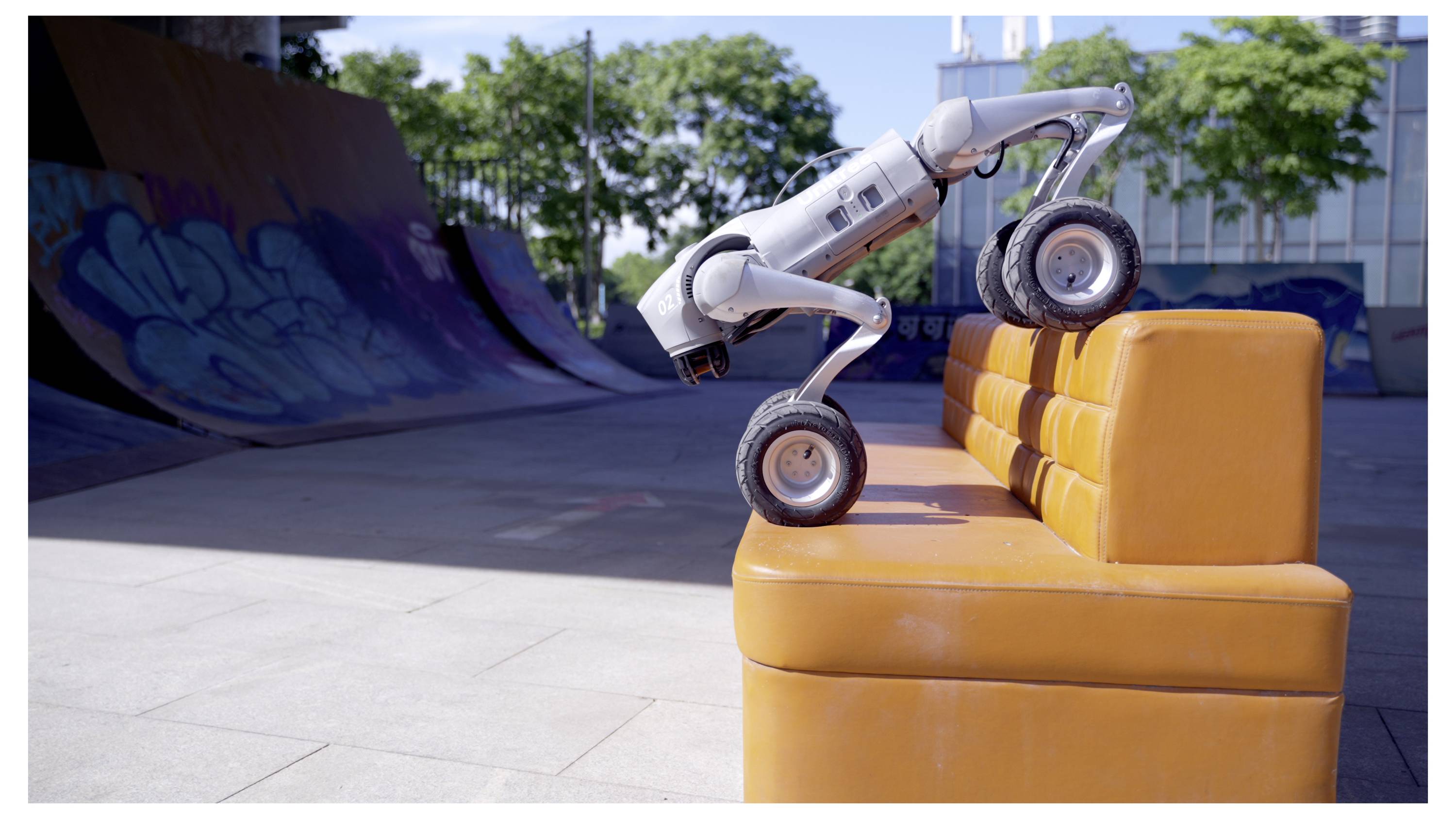 Ein Roboterhund klettert auf ein orangefarbenes Außensofa in einem Skatepark, mit einer Betonrampe und einer Graffiti-Wand im Hintergrund.