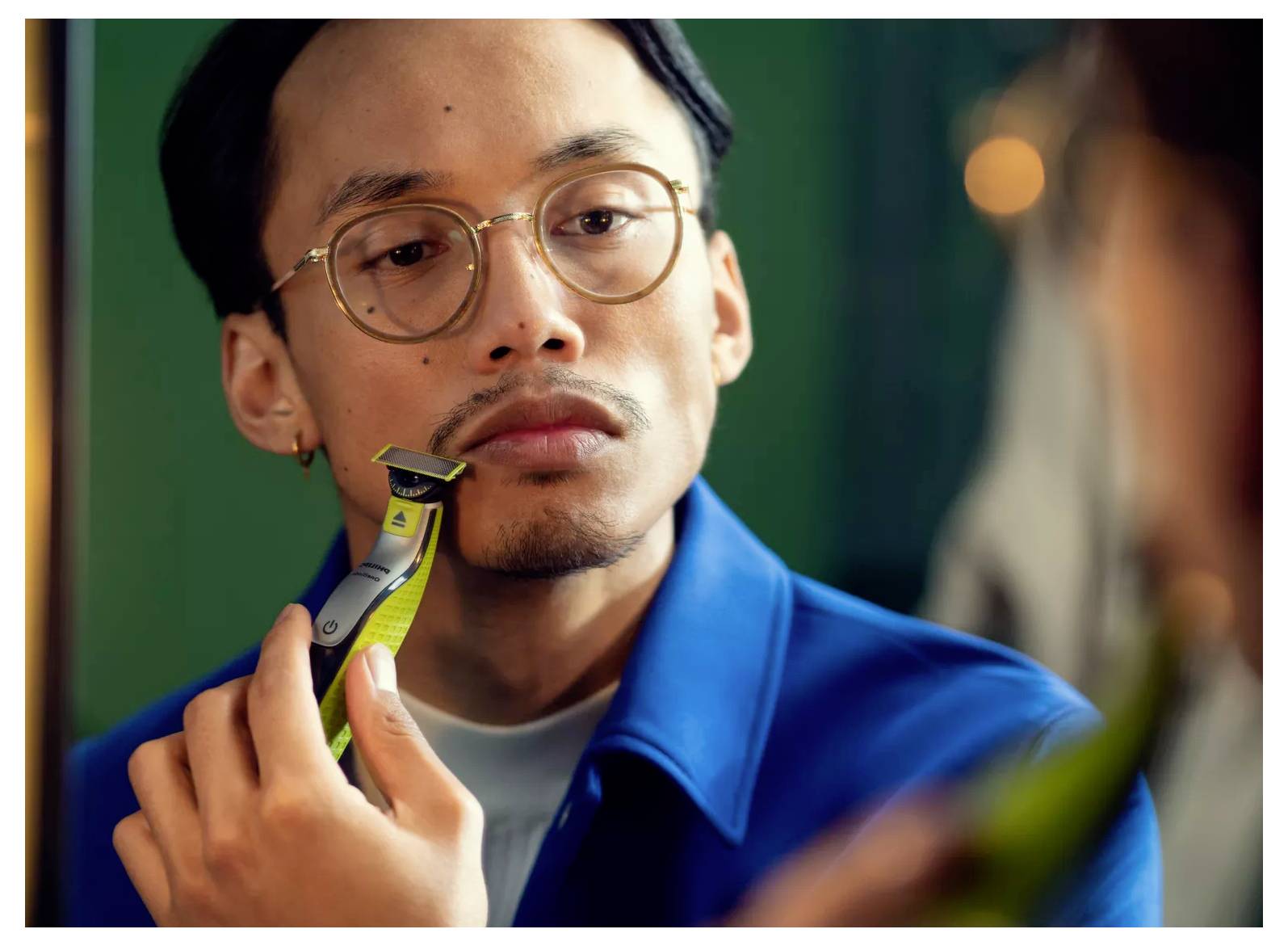 Ein Mann trimmt sich den Gesichtsbehang mit einem elektrischen Trimmer, während er sich in einem Spiegel anschaut, eine Brille trägt und ein blaues Hemd anhat.