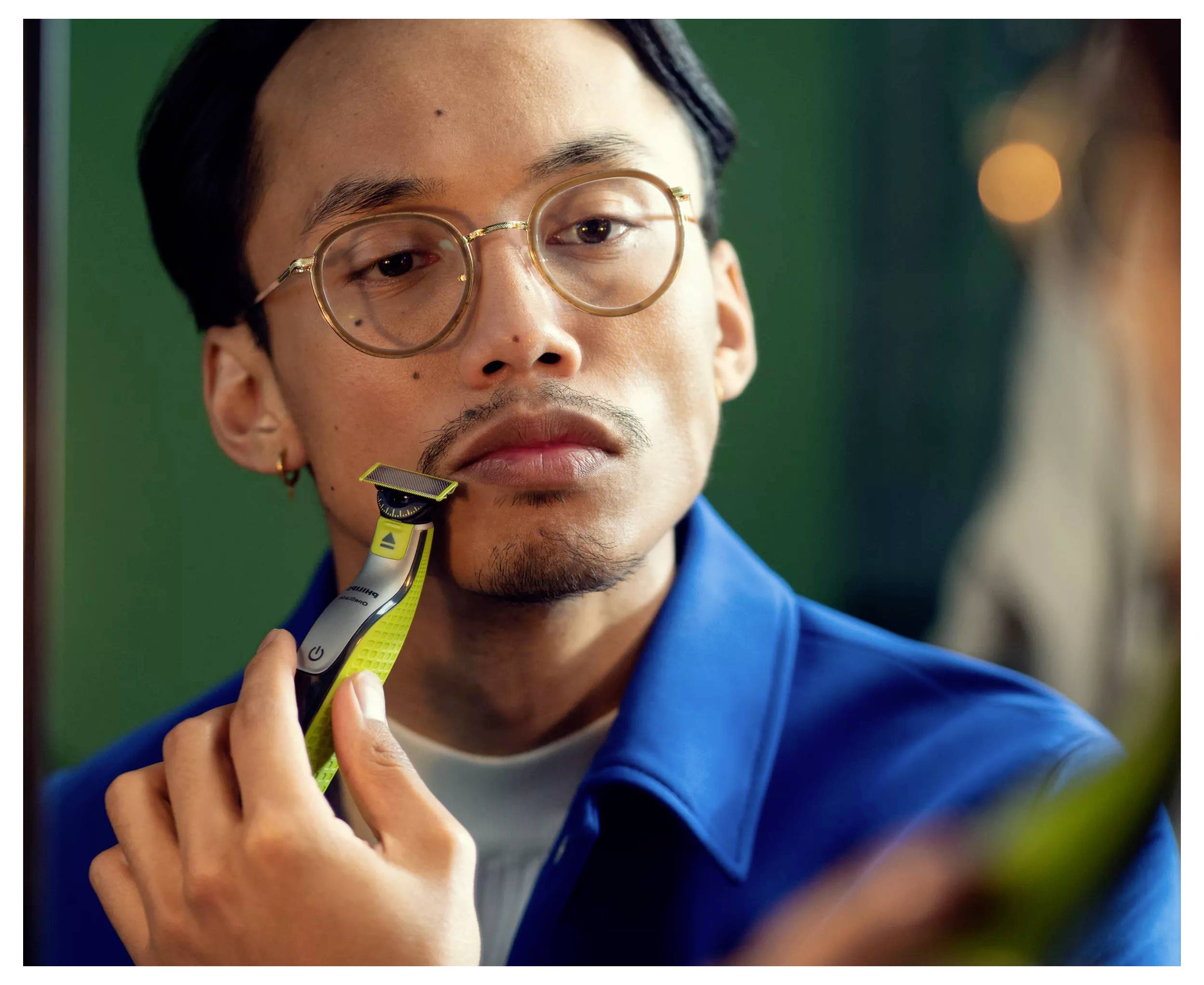 Ein Mann trimmt seinen Schnurrbart mit einem elektrischen Trimmer, während er in einen Spiegel schaut, trägt eine Brille und ein blaues Hemd.