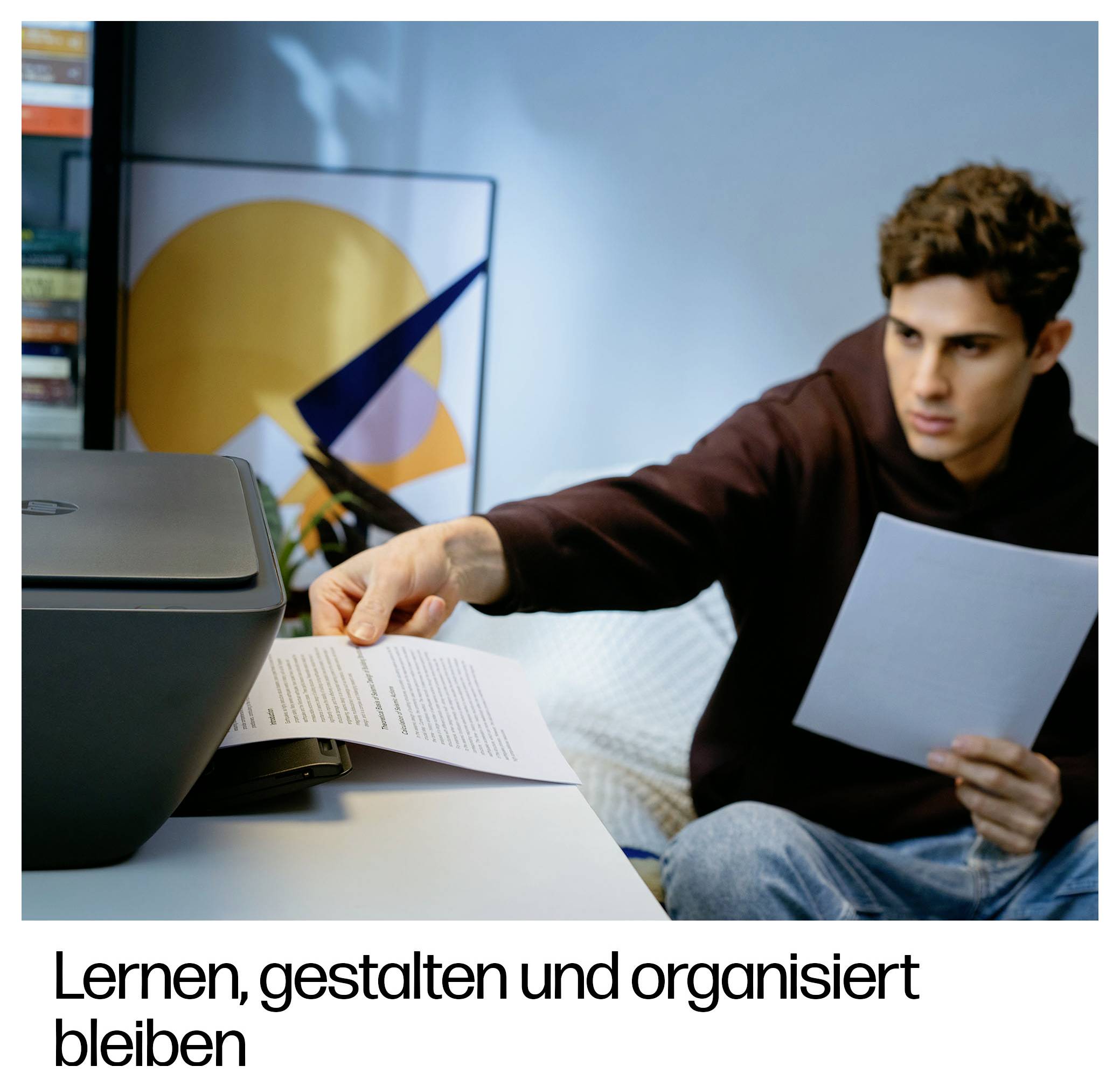 Junger Mann an einem Schreibtisch greift nach einer gedruckten Seite, während er eine andere hält, neben einem Drucker; deutscher Text liest sich „Lernen, gestalten und organisiert bleiben