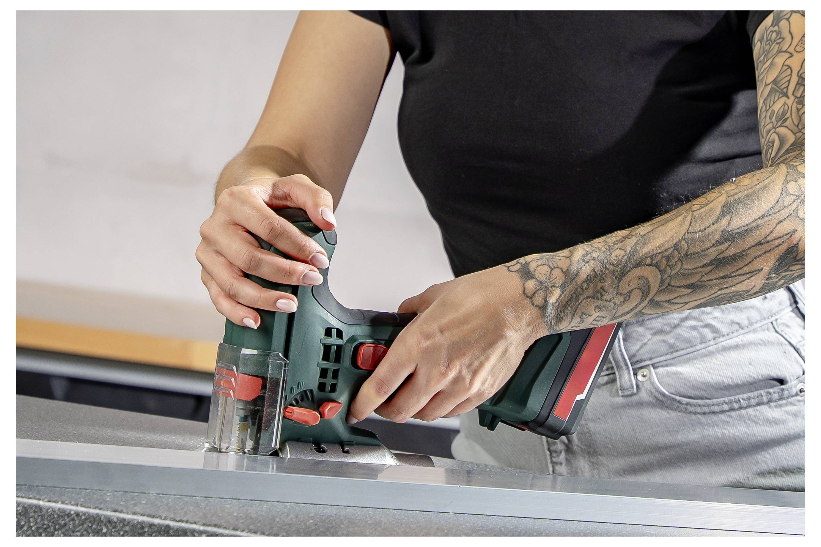 Person, die eine kabellose Stichsäge verwendet, um ein Material auf einer Werkbank zu schneiden, mit sichtbarem tätowiertem Arm.