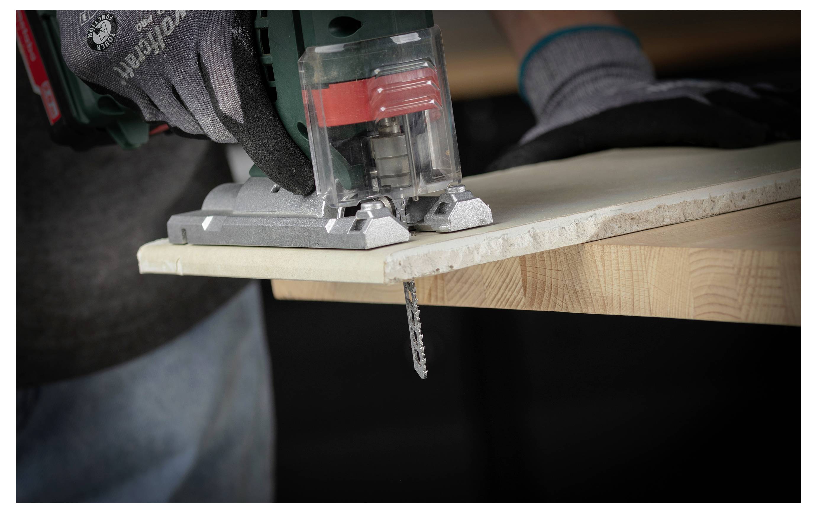 Eine handschuhtragende Person schneidet ein Holzbrett mit einer Stichsäge, wobei das Sägeblatt über die Kante des Bretts hinausragt.