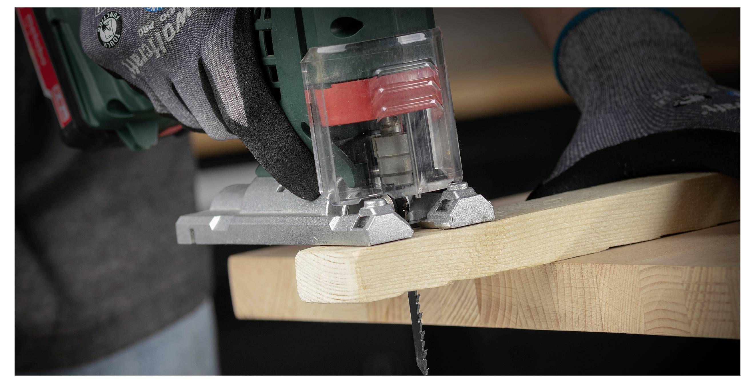 Nahaufnahme einer Person, die mit einer Stichsäge ein Holzbrett zuschneidet, mit behandschuhten Händen, die das Werkzeug führen.