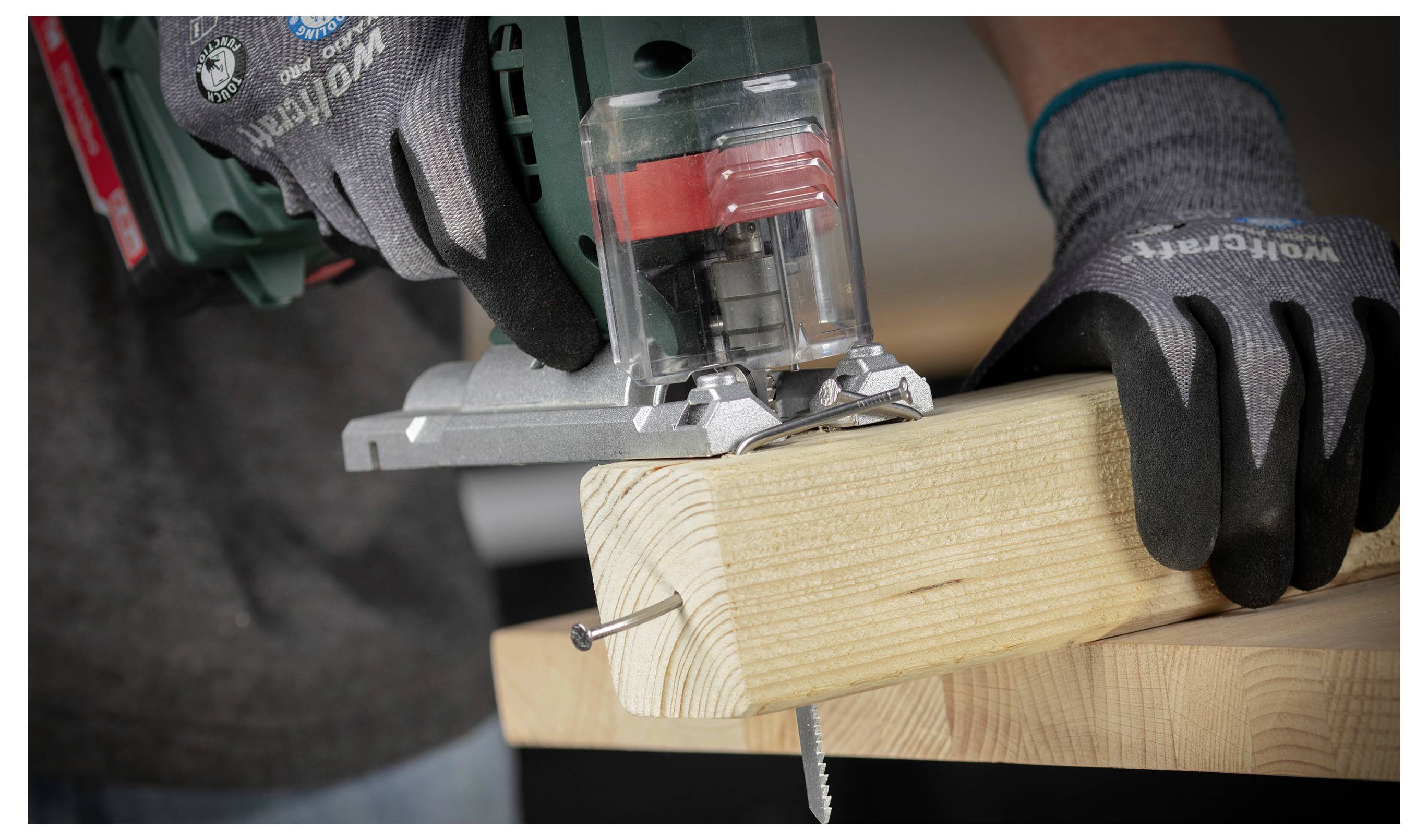 Handschuhe tragend, schneidet eine Person mit einer Elektrostichsäge ein mit Schrauben eingespanntes Holzbrett in einer Werkstatt.