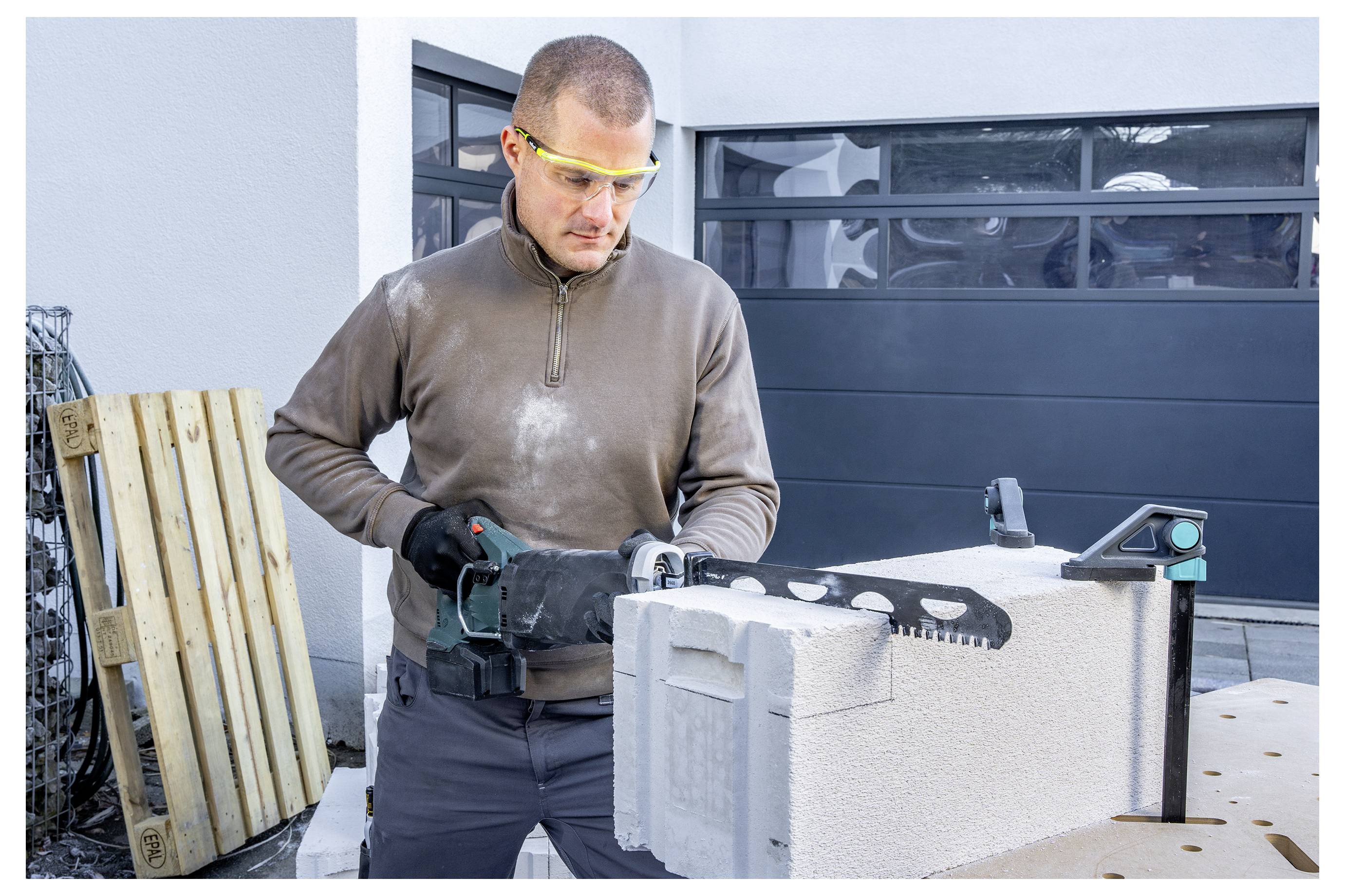 Ein Mann schneidet einen Betonblock mit einer tragbaren Elektrosäge im Freien auf einer Einfahrt, wobei er eine Schutzbrille und Handschuhe trägt.
