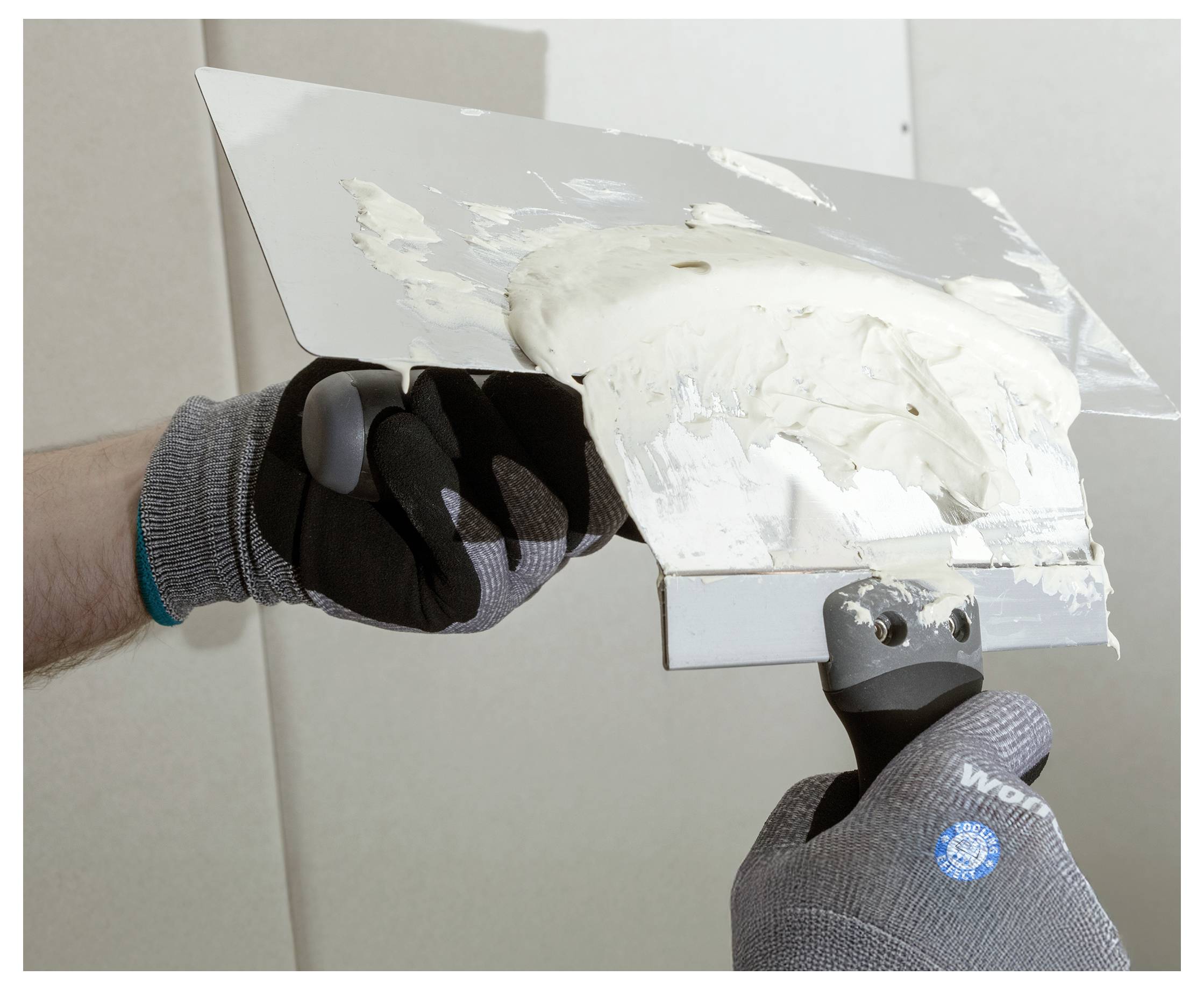 Behandschuhte Hände halten ein Metallspachtel, der mit weißem Trockenbauspachtel bedeckt ist und zeigen eine Wandreparatur- oder Verputzaufgabe.