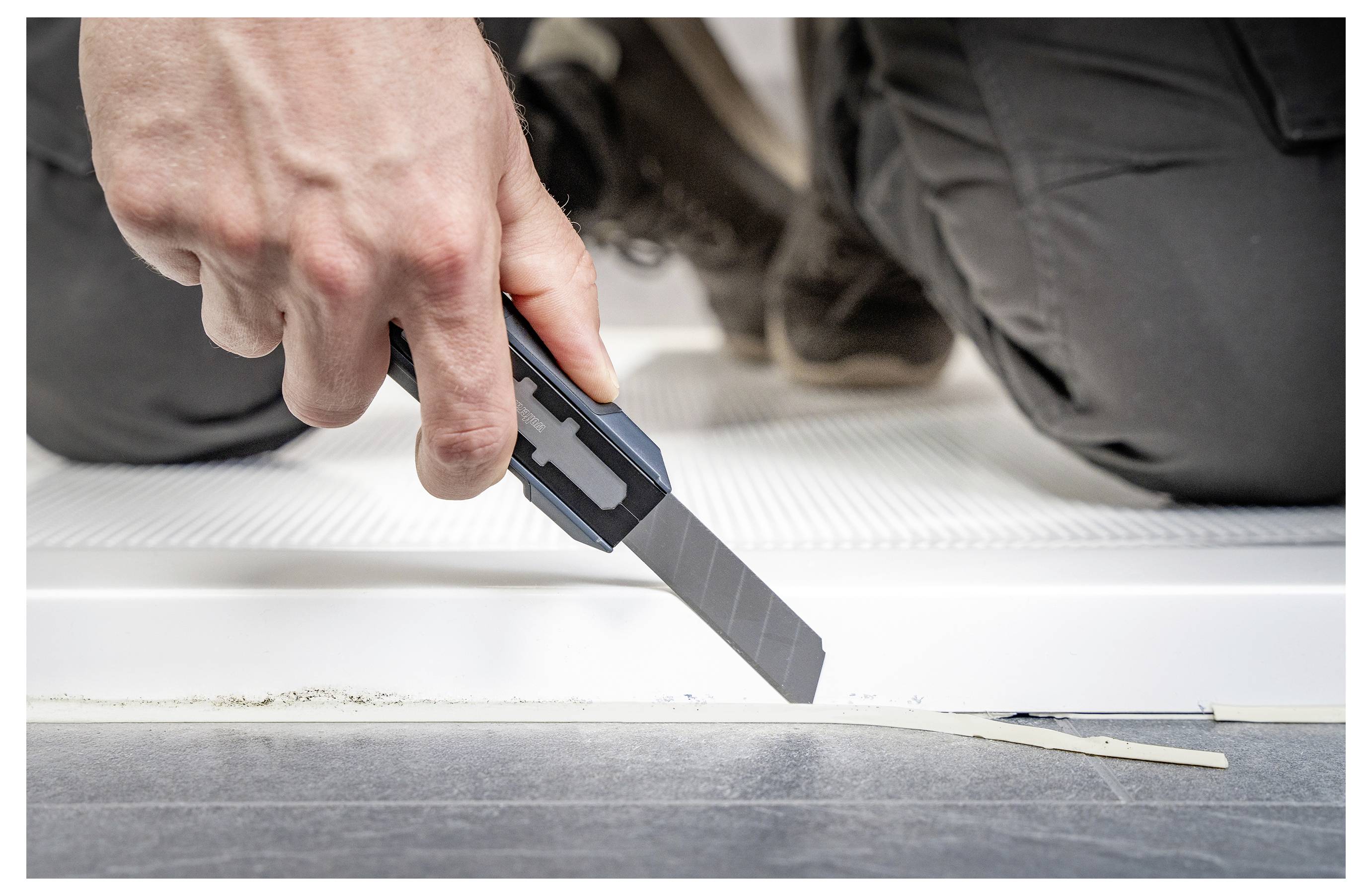 Nahaufnahme einer Hand, die ein Universalmesser verwendet, um Material entlang der Kante eines Bodens oder einer Sockelleiste während der Installation oder Reparatur zu schneiden oder zu trimmen.