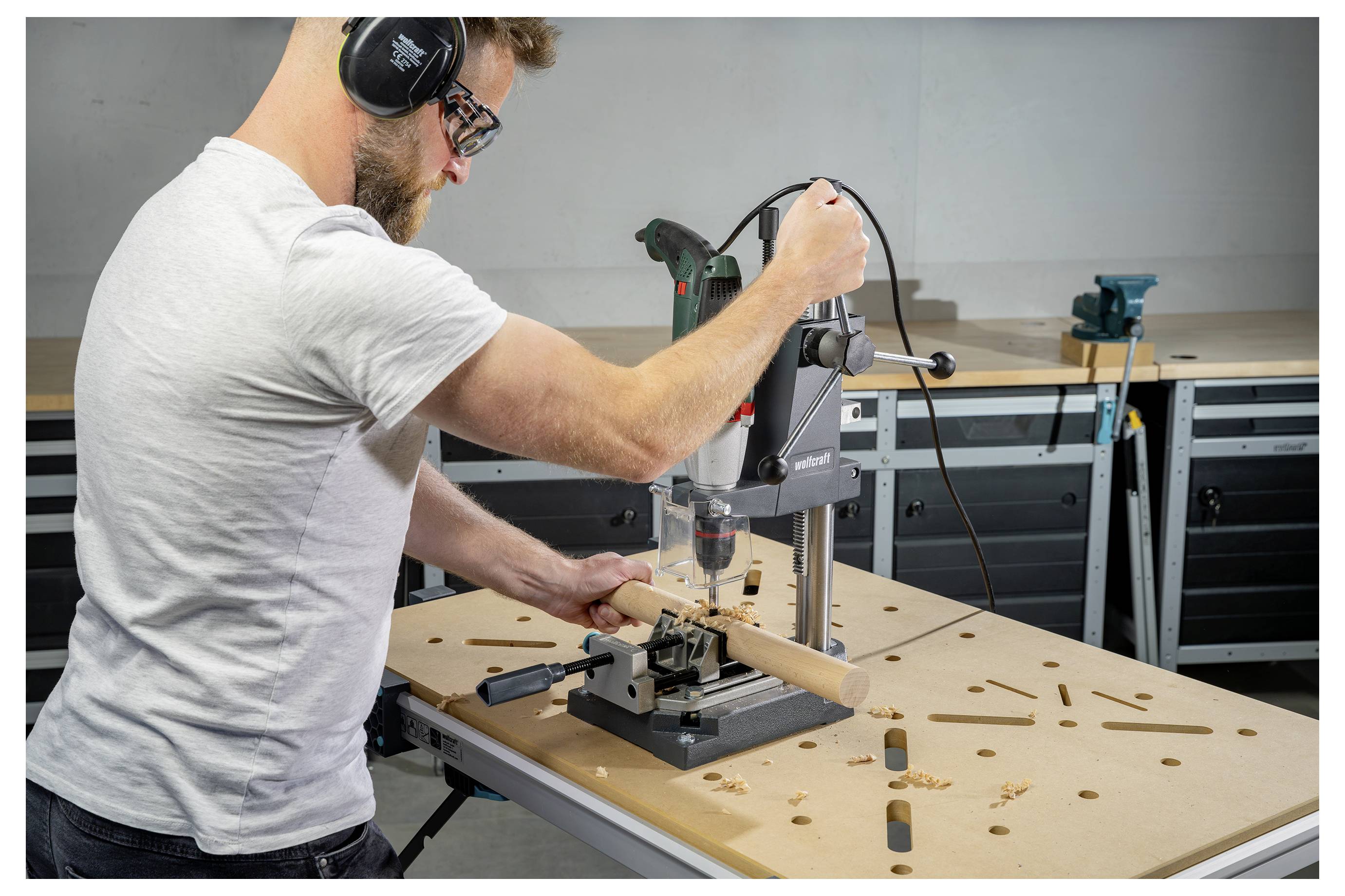Ein Mann benutzt eine Bohrmaschine, um ein Loch in einen Holzdübel auf einer Werkbank in einer Werkstatt zu bohren, mit Spannzwingen und Werkzeugen in der Nähe.
