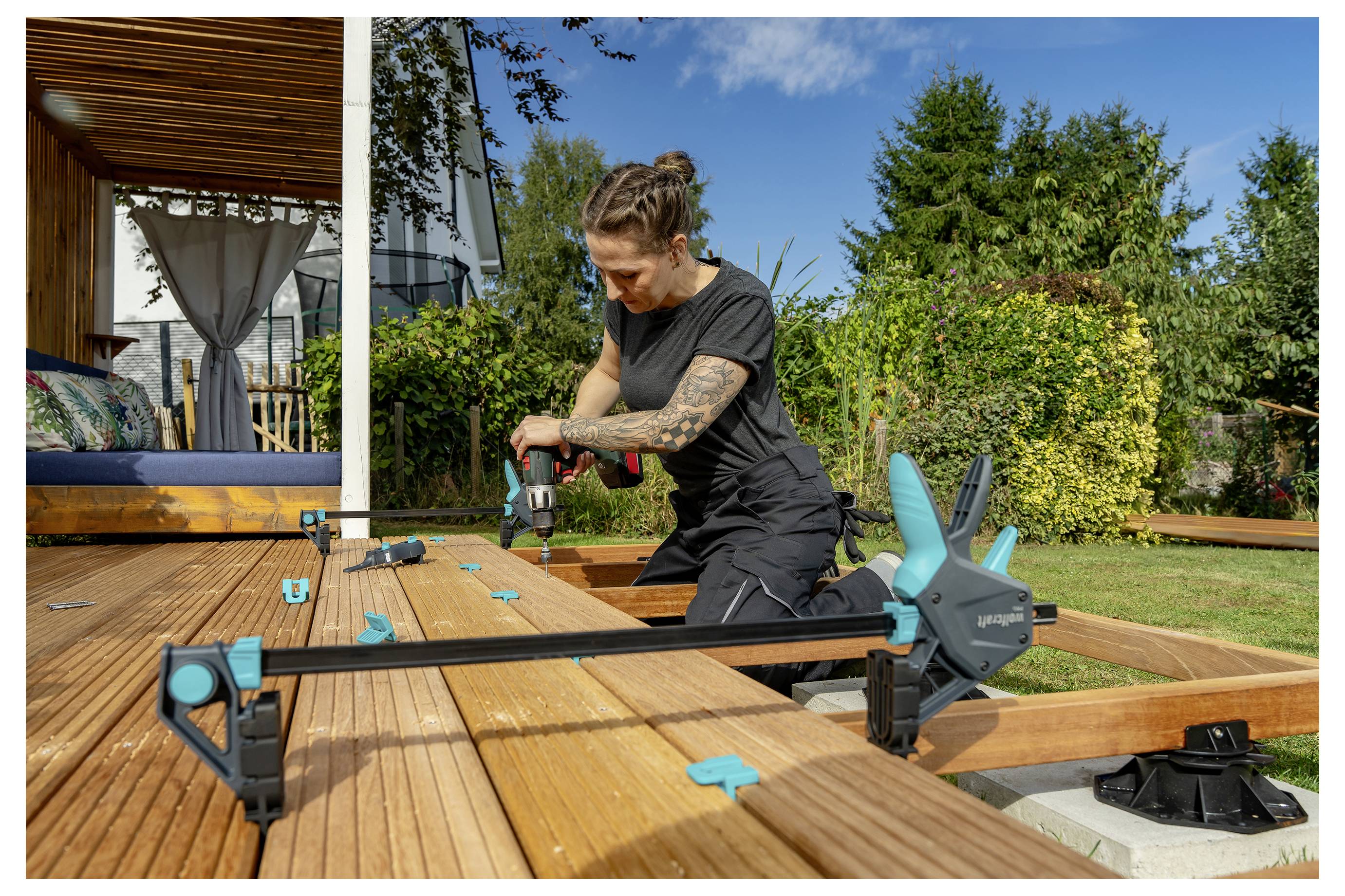 Frau, die einen Bohrer verwendet, um einen Holzdeckrahmen an einem sonnigen Tag im Freien zusammenzubauen.