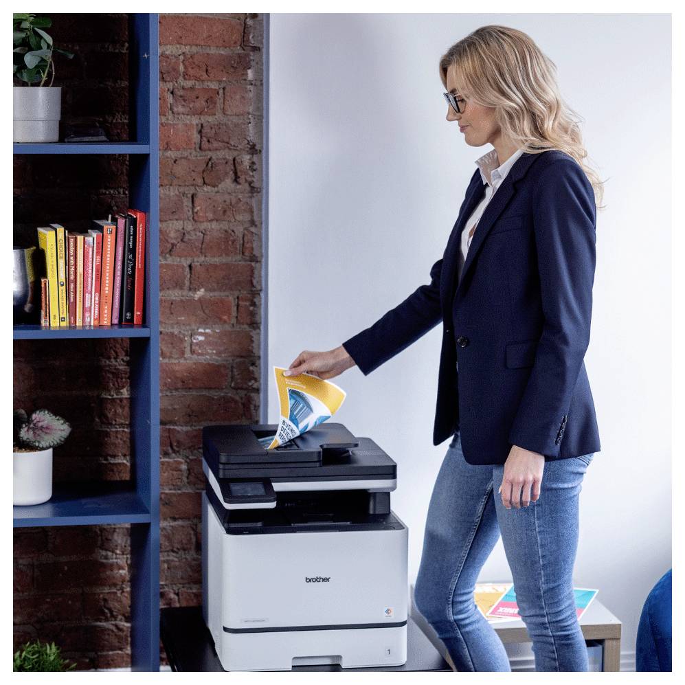 Eine Frau in einem Blazer legt ein Blatt in einen Brother-Drucker neben einem Bücherregal in einem modernen Büro- oder Wohnarbeitsbereich ein.