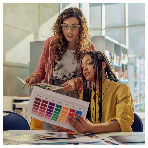 Zwei Frauen überprüfen gedruckte Farbmuster und Designentwürfe an einem Tisch und besprechen ein Projekt in einem hellen Studio oder Klassenzimmer.