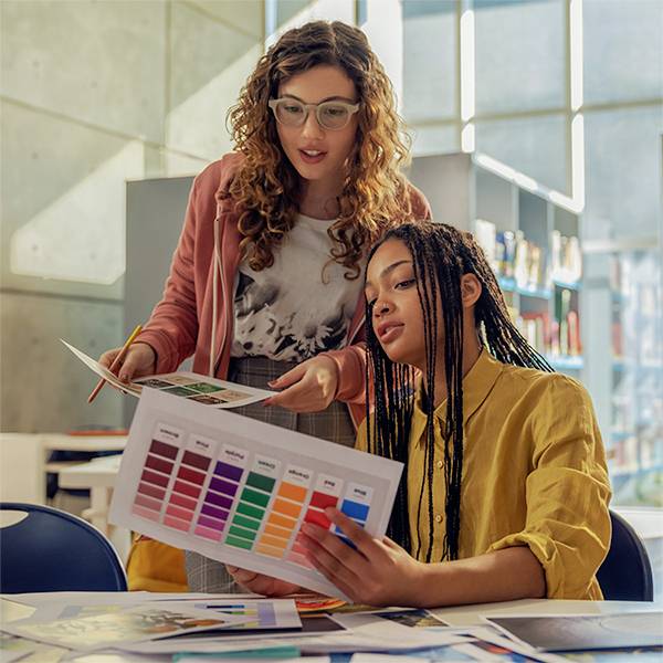 Zwei Frauen betrachten zusammen gedruckte Farbmuster und Designmuster in einem hellen Klassenzimmer oder Studio.