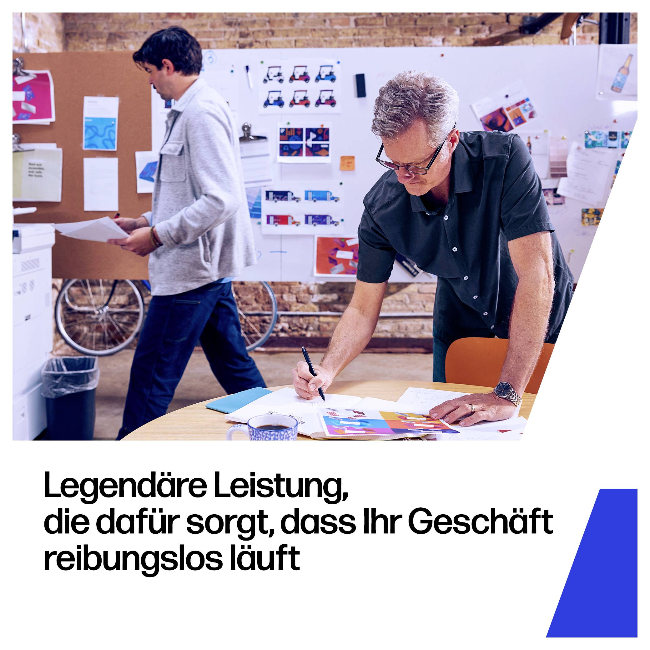 Zwei Männer arbeiten in einem Bürostudio mit Designboards; einer schreibt an einem Tisch, während der andere eine Kiste trägt. Deutsche Überschrift: 'Legendäre Leistung, die dafür sorgt, dass Ihr Geschäft reibungslos läuft'
