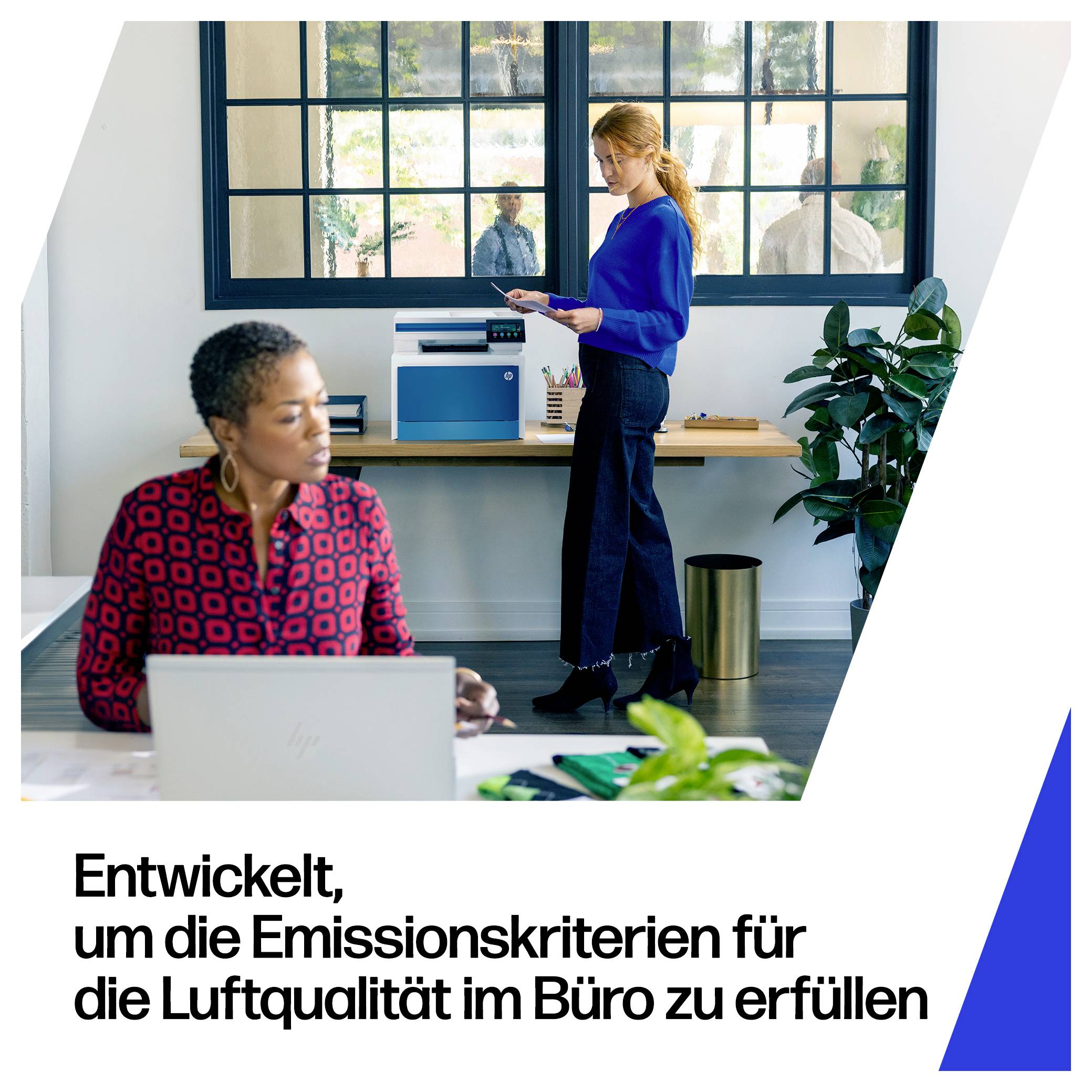 Zwei Frauen in einem modernen Büro, eine arbeitet an einem Laptop und eine hält Papiere in der Nähe eines Schreibtisches, mit deutschem Text, der lautet: „Entwickelt, um die Emissionskriterien für die Luftqualität im Büro zu erfüllen
