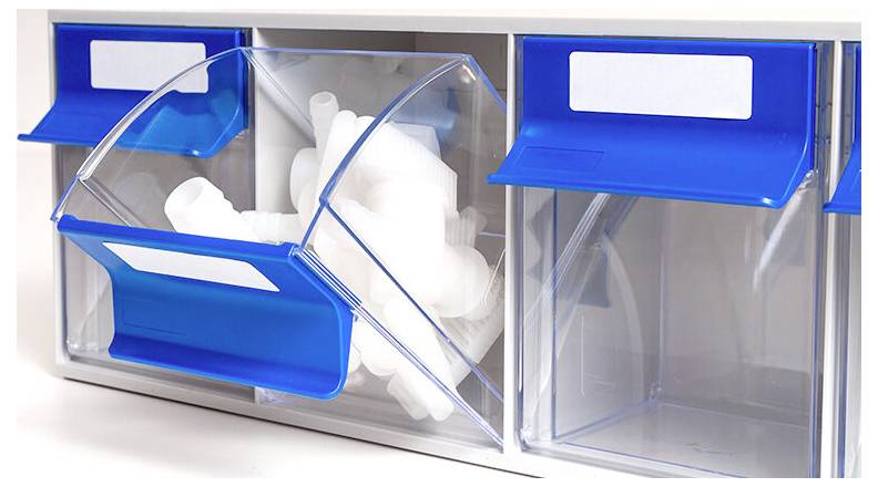 An der Wand montierter transparenter Kunststofforganizer mit blauen Scharnierdeckeln, teilweise geöffnet und gefüllt mit weißen Wattestäbchen oder Wattepads.