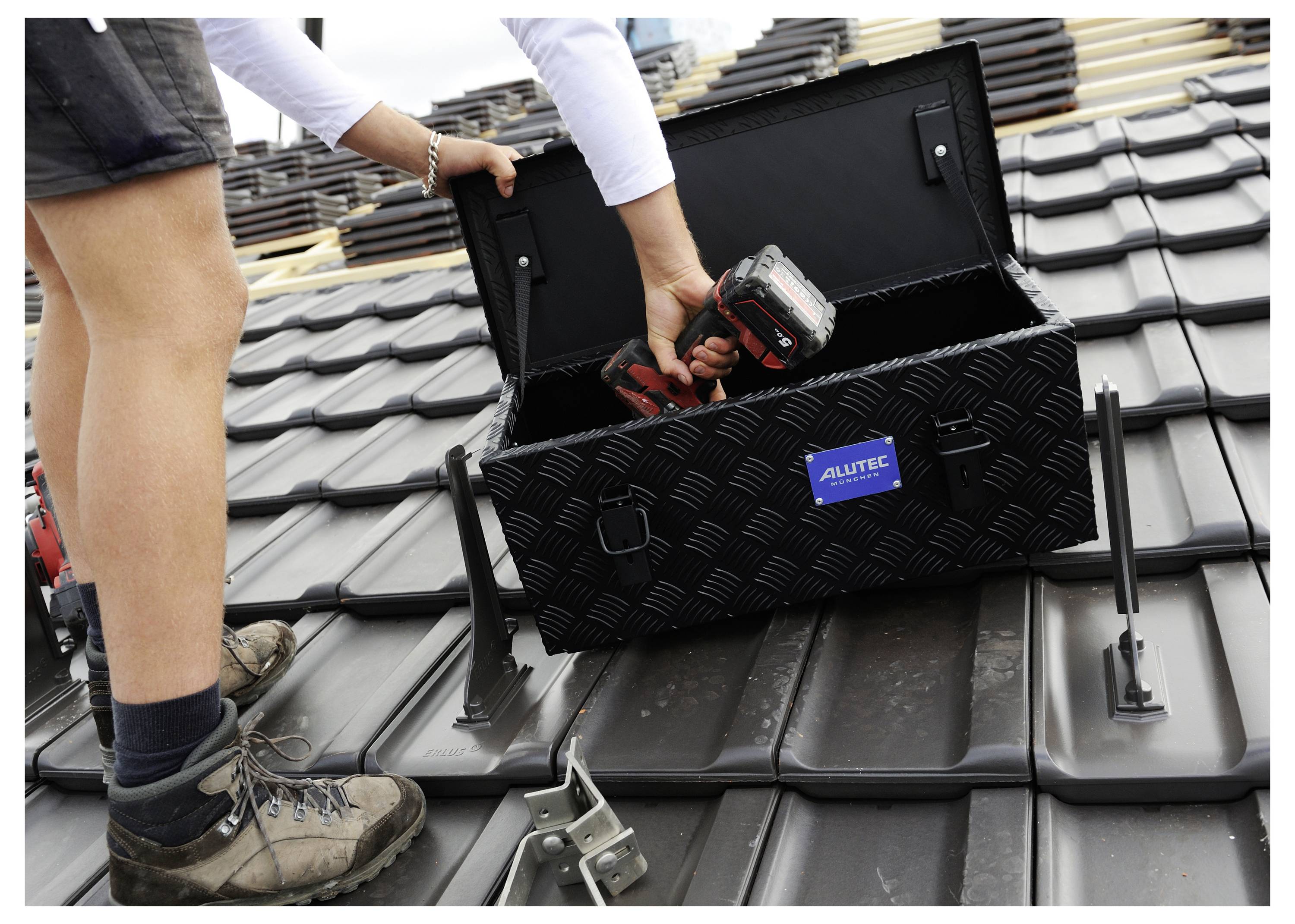 Person auf einem Dach installiert eine schwarze Riffelblech-Dachlagerbox, während sie eine kabellose Bohrmaschine hält, wobei Dachziegel und Befestigungshardware sichtbar sind.