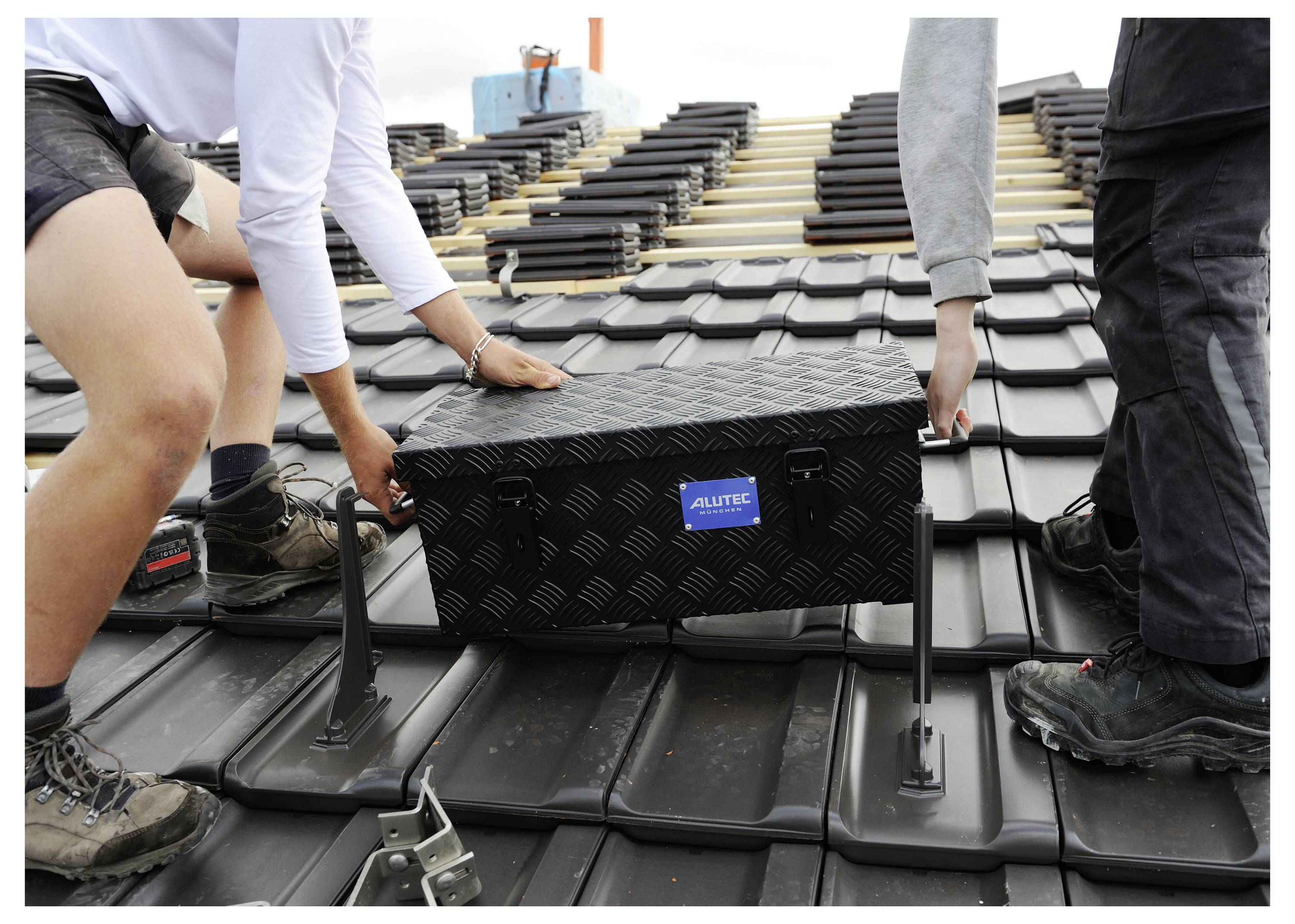 Zwei Arbeiter installieren eine schwarze Dachmontagebox auf einem Ziegeldach, mit gestapelten Materialien im Hintergrund.