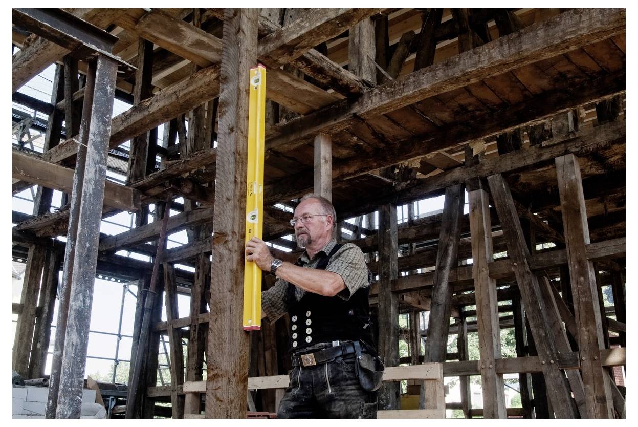 Ein Mann in Arbeitskleidung verwendet eine Wasserwaage, um einen Holzbalken in einer teilweise ausgebauten, alten Holzständerkonstruktion zu prüfen.