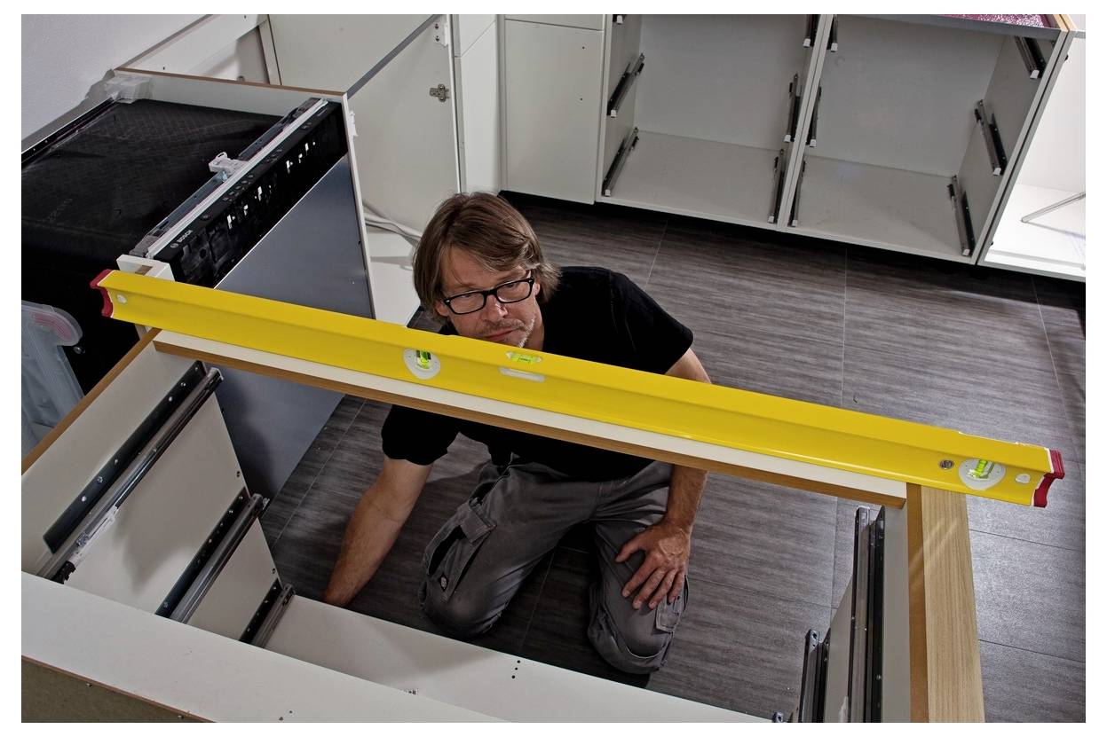 Ein Mann kniet im Inneren einer Kücheneinbauschrank-Installation und prüft die Ausrichtung mit einer gelben Wasserwaage über den Arbeitsplattenrahmen.