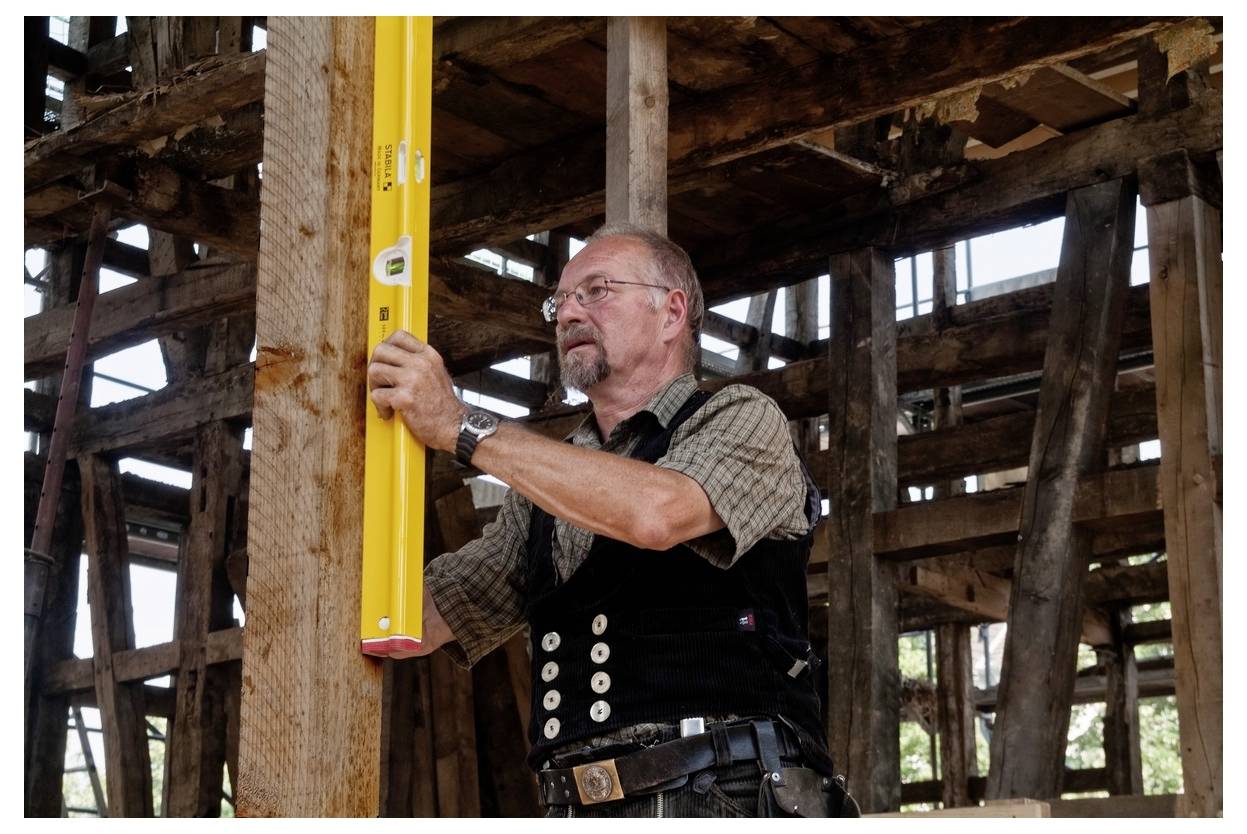 Bauarbeiter, der mit einer gelben Wasserwaage einen Holzpfosten in einem sich im Bau befindlichen Fachwerkgebäude überprüft.