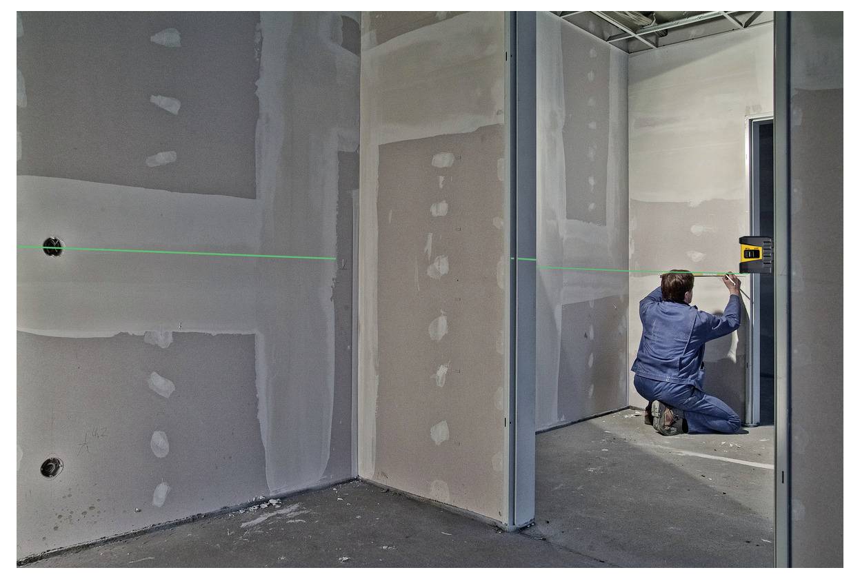 Arbeiter in blauem Schutzanzug kniet in einem unfertigen Raum und misst eine Wand mit einer Laserwasserwage während der Innenausbau.