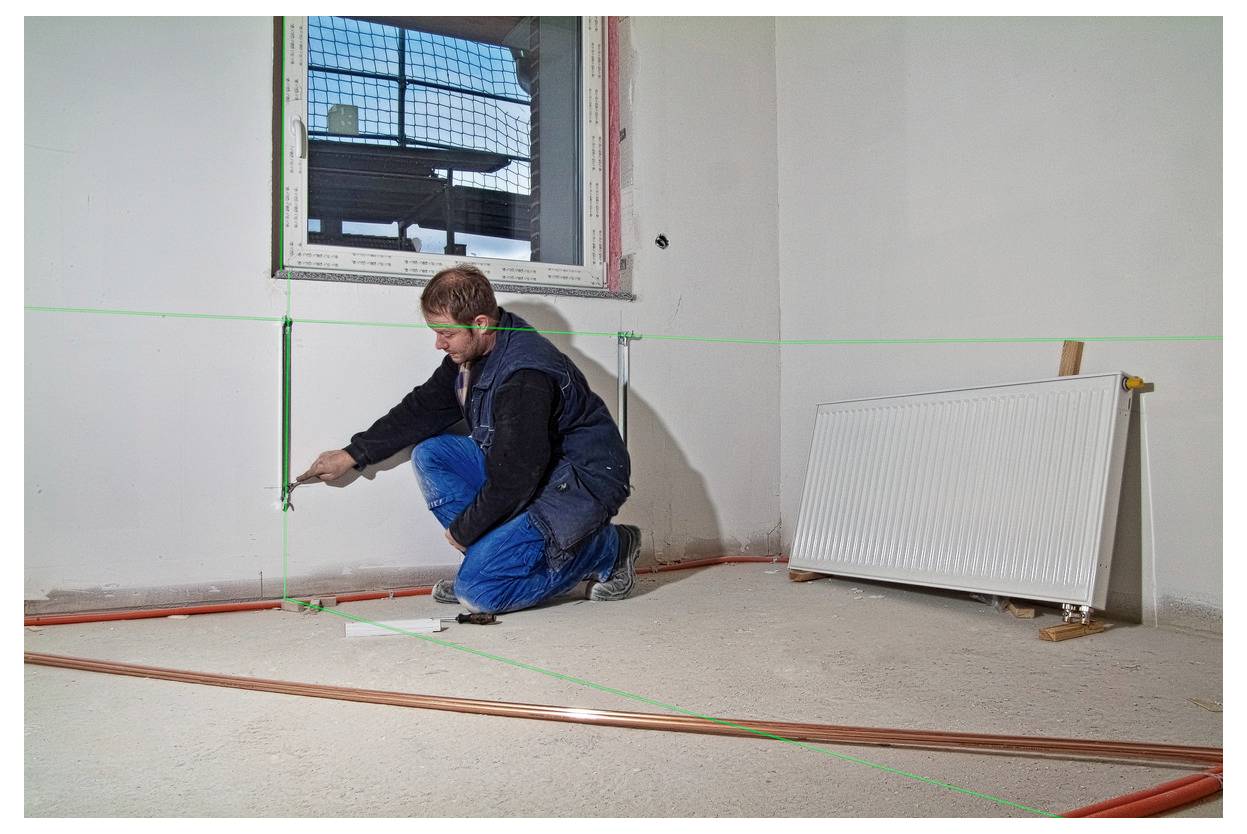 Arbeiter hockt sich in einem unfertigen Zimmer hin und misst oder richtet Wandrohrleitung in der Nähe eines Fensters und eines an der Wand lehnenden Heizkörpers aus.