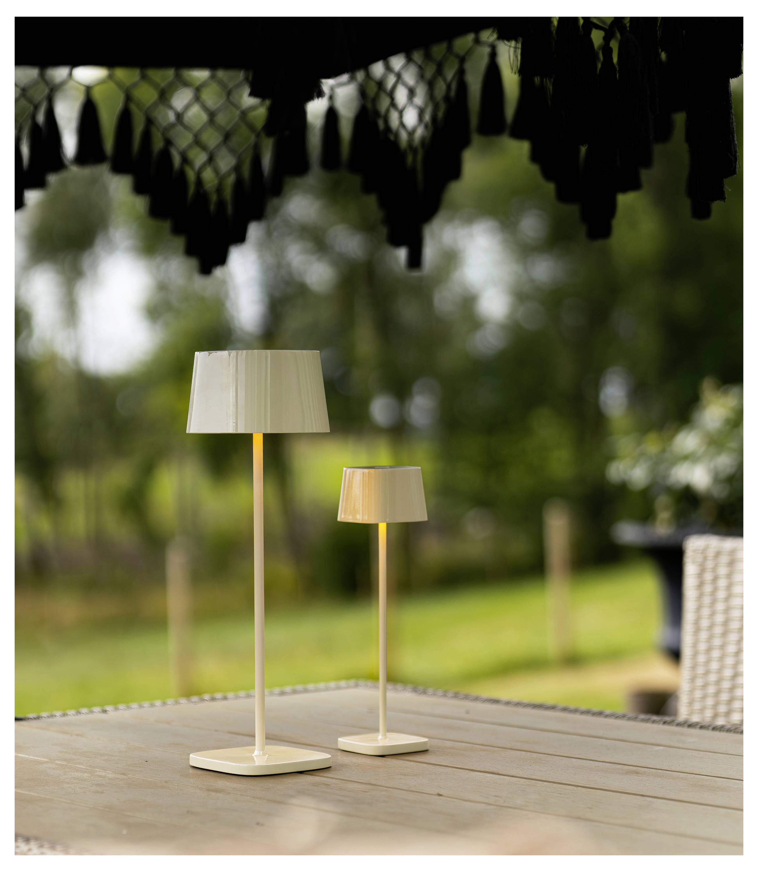 Zwei kleine cremefarbene Tischlampen auf einem Holztisch im Freien unter einer schattigen Pergola, mit einem unscharfen Garten im Hintergrund.