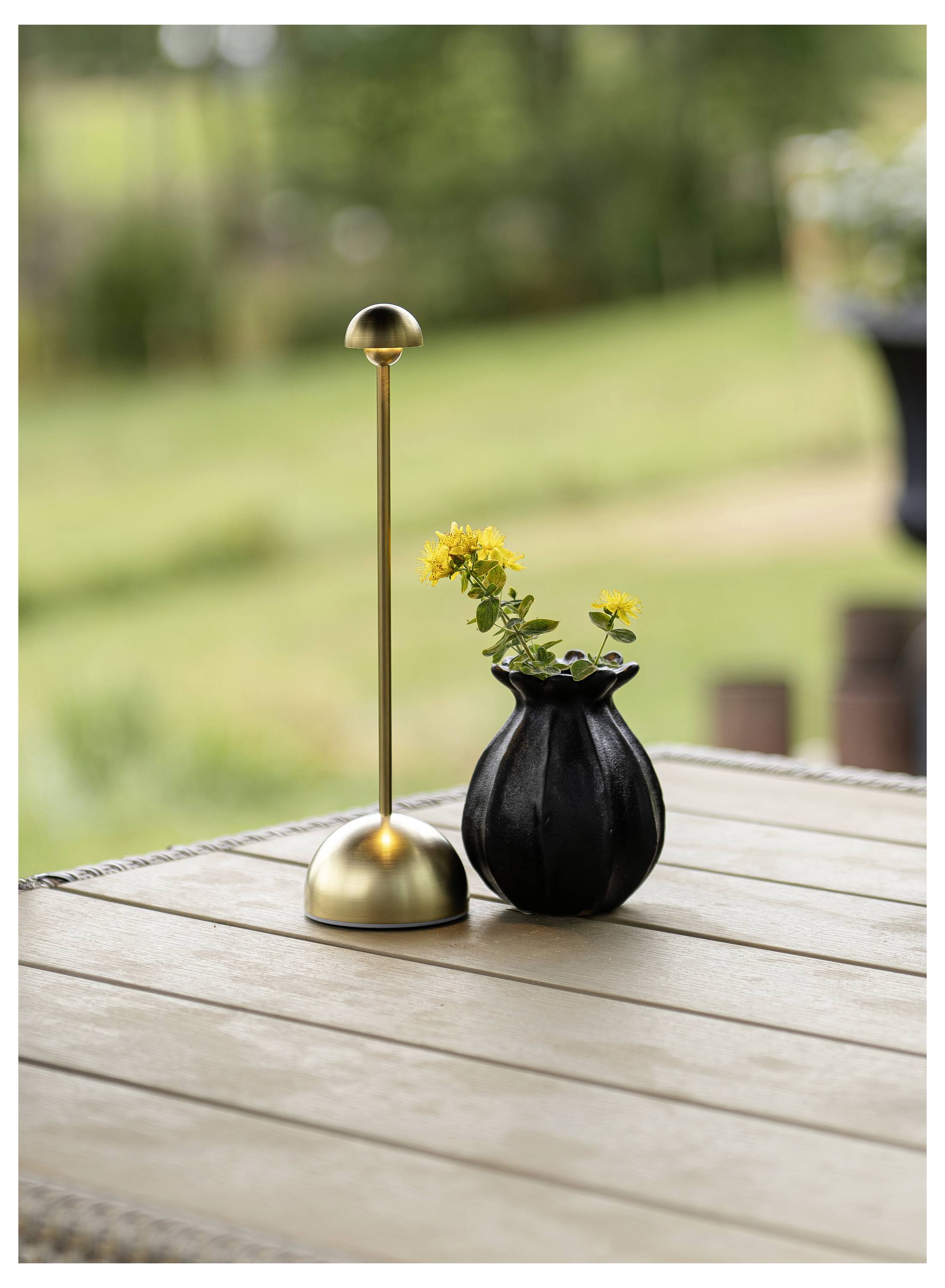 Eine goldene Tischlampe und eine kleine schwarze Vase mit gelben Blumen stehen auf einem hölzernen Außentisch, mit unscharfem Gartenhintergrund.