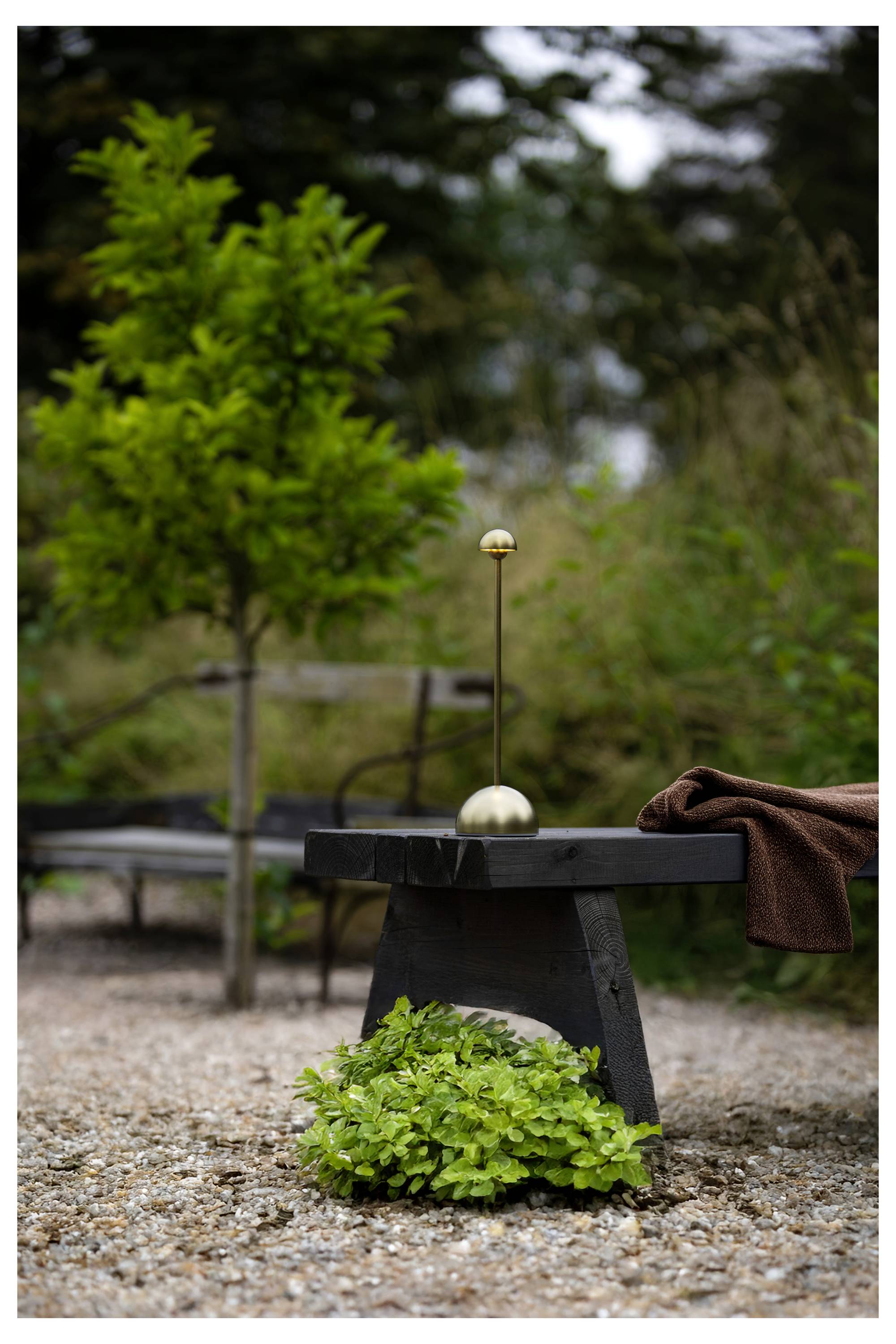 Outdoor-Patio-Beistelltisch mit einer messingfarbenen lampenähnlichen Mittelpunktfigur und einem braunen Handtuch, das darüber drapiert ist, umgeben von Grün und Kies.