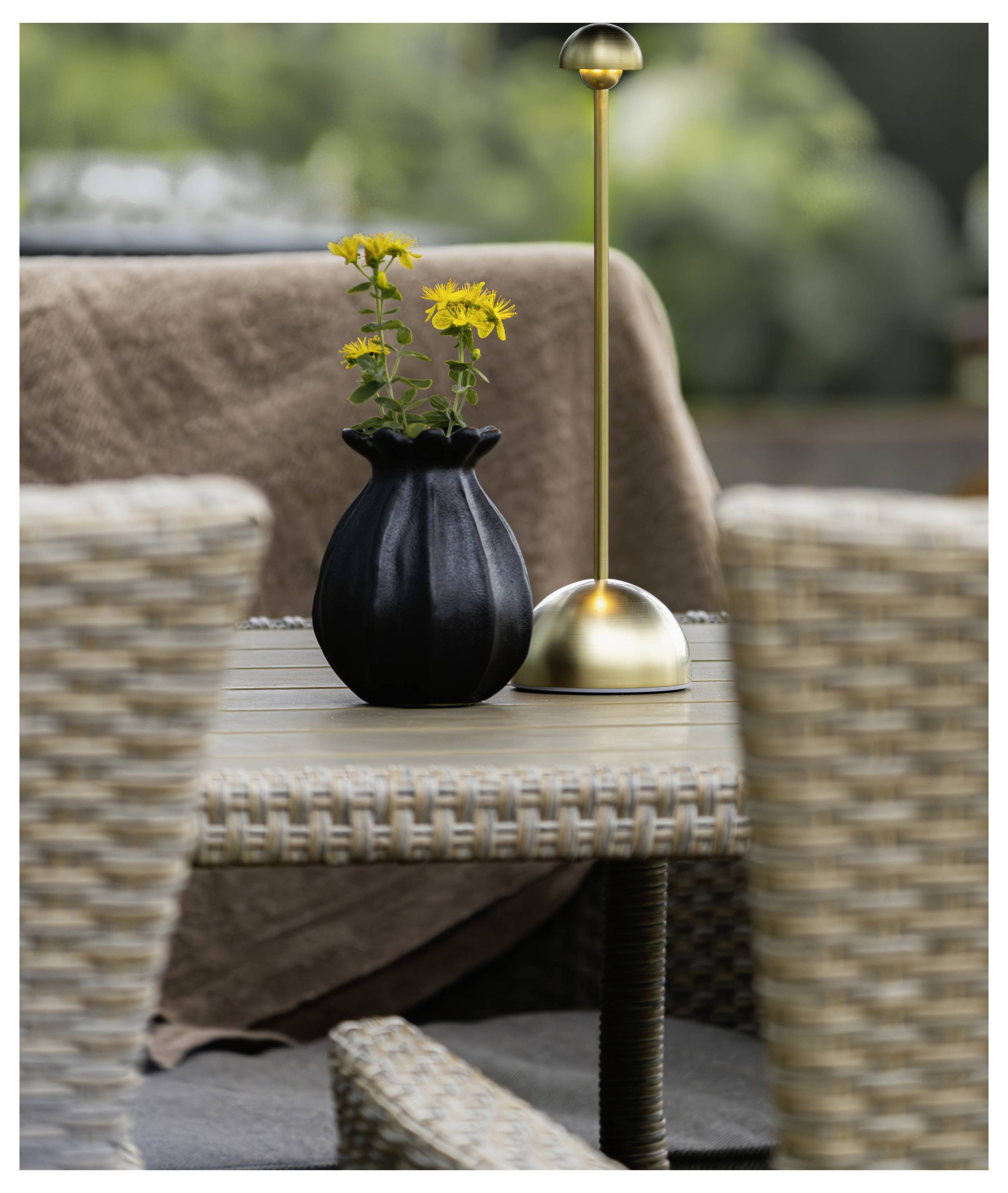 Schwarze Vase mit gelben Blumen und einer Messingtischleuchte auf einem geflochtenen Outdoor-Tisch, mit Korbstühlen und einer braunen Wurfdecke im Hintergrund.