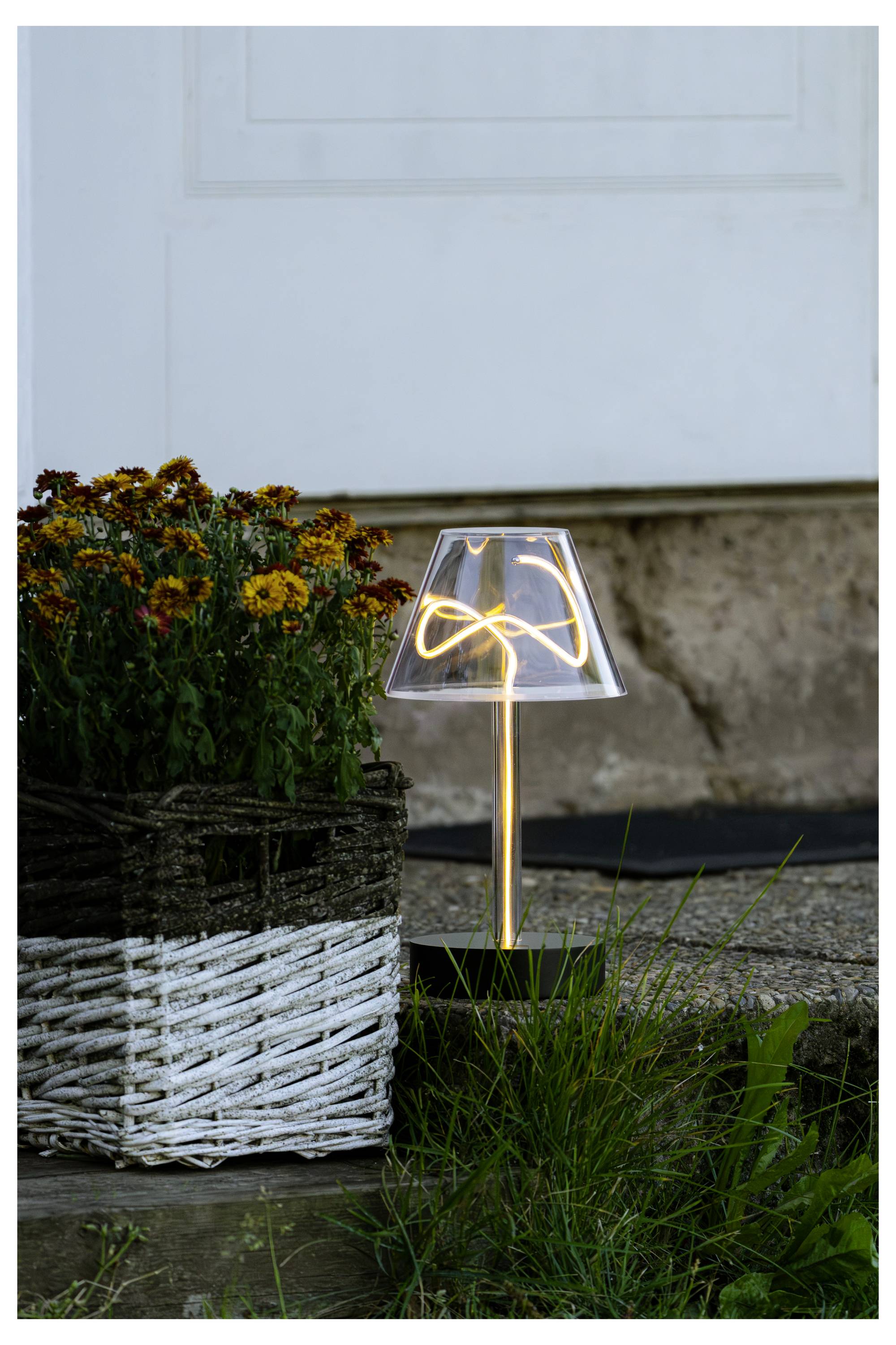 Kleine leuchtende Tischlampe neben einem weißen Korbblumenkübel mit Blumen und Gras, auf einer Türschwelle vor einer weißen Tür aufgestellt.