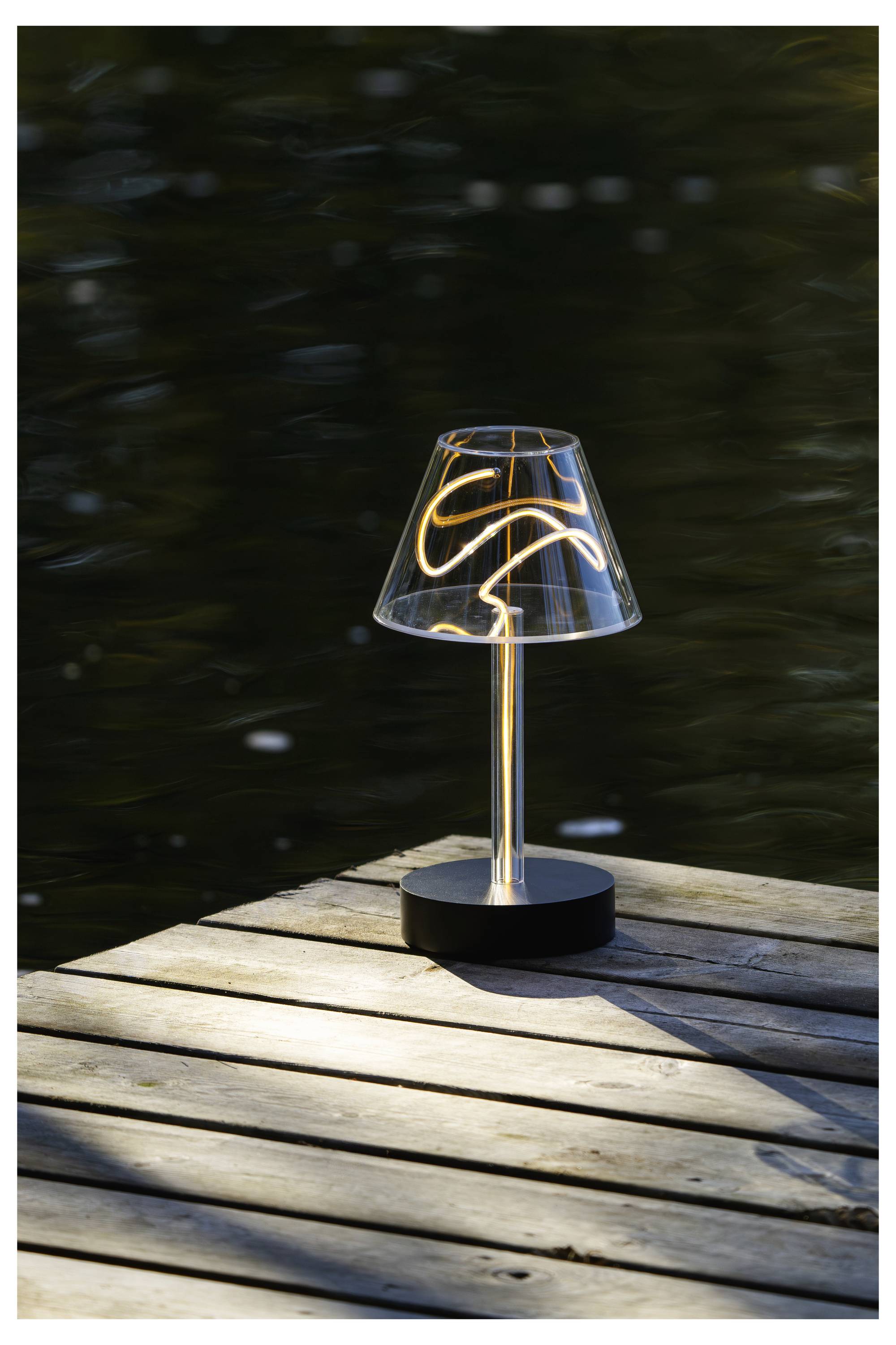 Eine kleine leuchtende Tischlampe auf einem Holzsteg neben dunklem Wasser, die bei Einbruch der Dämmerung Licht auf die Dielen wirft.