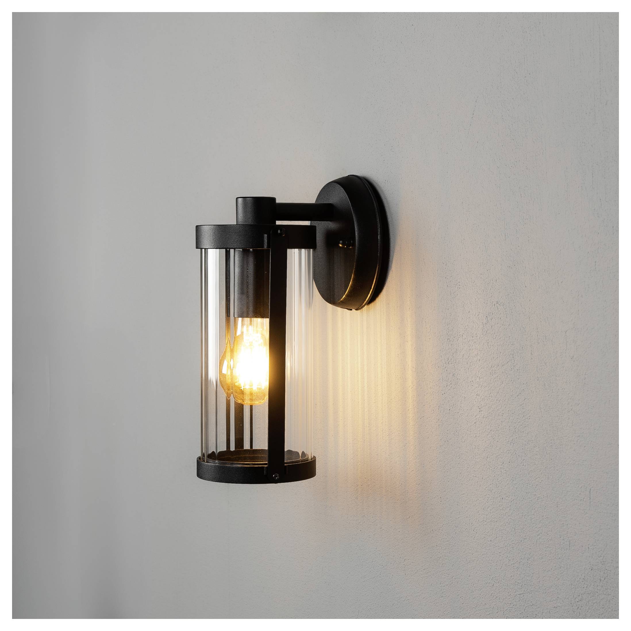 Schwarze Wandleuchte mit freiliegender leuchtender Glühbirne wirft warmes Licht und vertikale Schattenlinien auf eine schlichte hellgraue Wand.