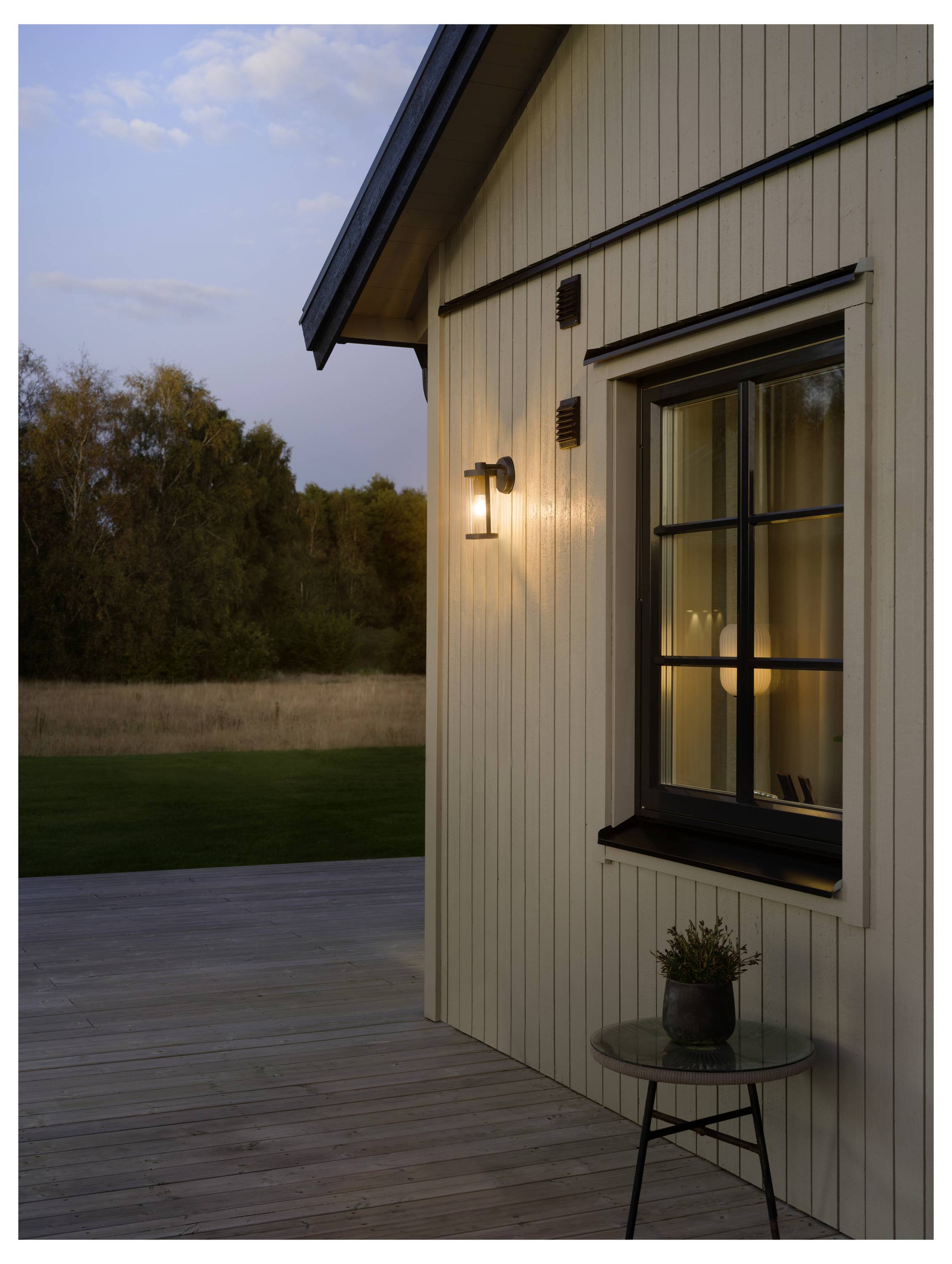 Außenseite eines modernen Hauses bei Einbruch der Dunkelheit, mit einer beleuchteten Wandleuchte neben einem Fenster und einem kleinen Tisch auf der Holzterrasse.