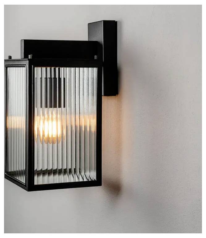 Moderne schwarze Wandleuchte mit geripptem Glasschirm, der warm auf einer hellgefärbten Wand leuchtet und einen sanften Schatten wirft.