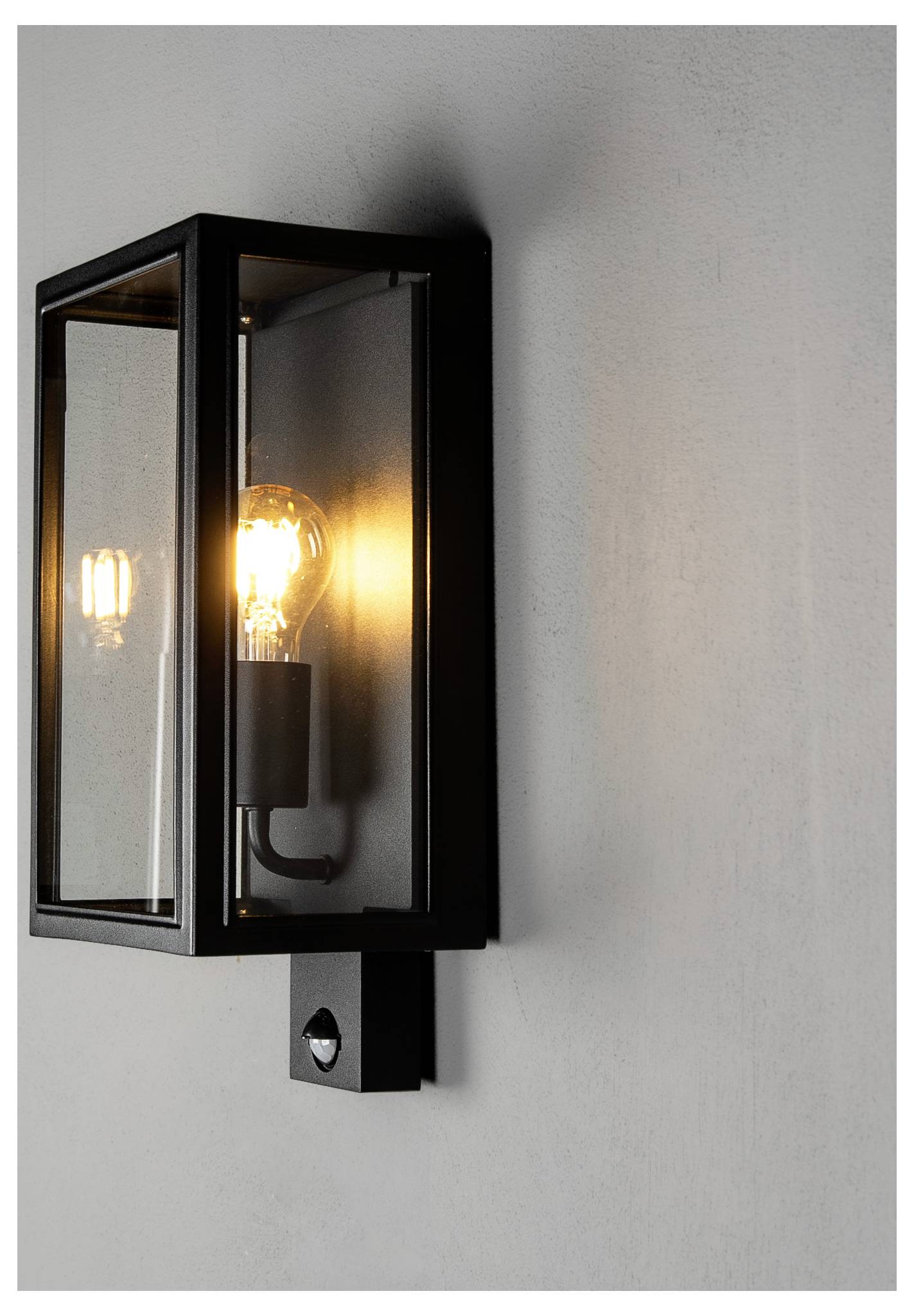 Schwarze Wandleuchte mit Glaspaneelen und einer leuchtenden Glühbirne, die an einer hellgrauen Wand montiert ist.