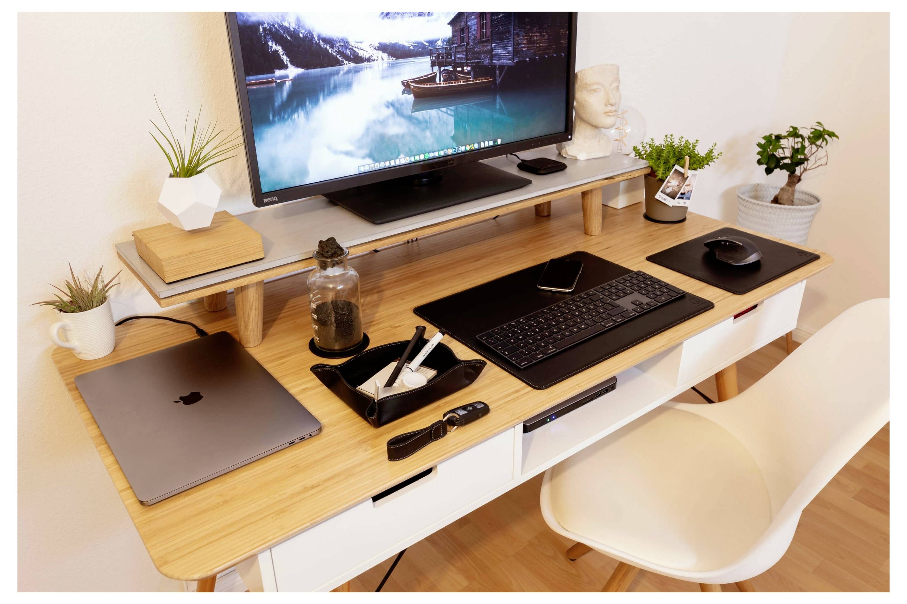Ein aufgeräumter Schreibtisch im Homeoffice mit einem Monitor, Laptop, Tastatur, Maus, Notizbuch, Stiftablage und kleinen Topfpflanzen.