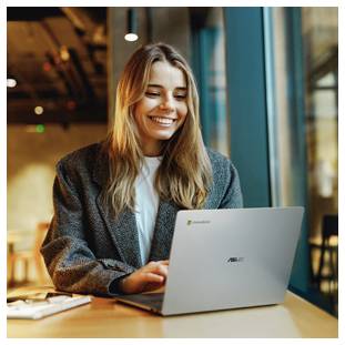 Lächelnde Frau, die an einem Laptopgerät an einem Caféatisch arbeitet, neben einem Fenster in einer zwanglosen Innenumgebung.