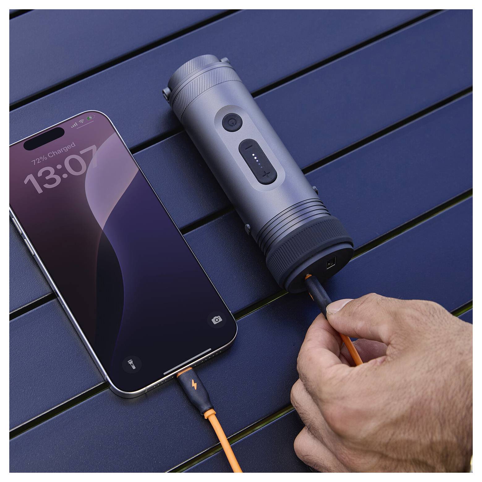 Hand steckt ein USB-C-Kabel in ein tragbares zylindrisches Ladegerät neben einem Smartphone, das auf einem dunklen Tisch aufgeladen wird.
