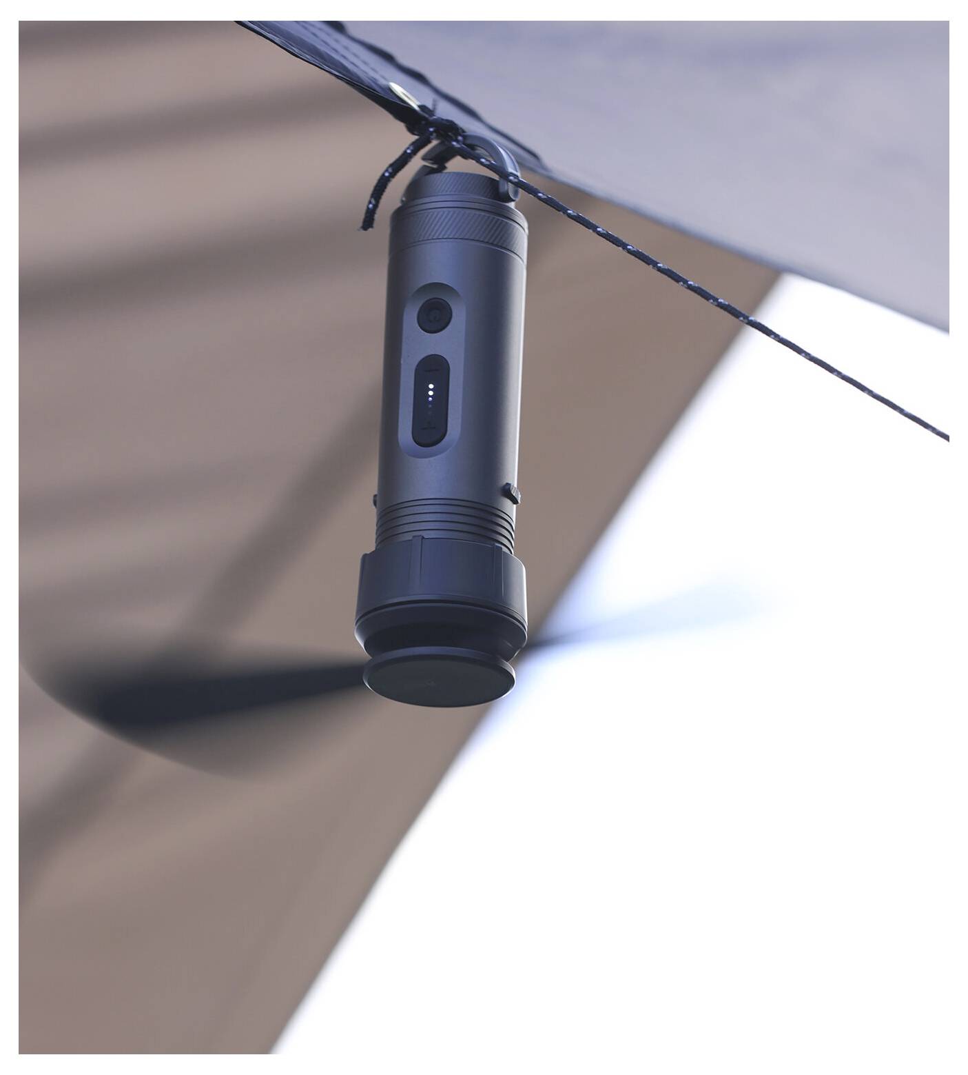 Nahaufnahme einer schwarzen zylindrischen Taschenlampe, die an einer Schnur unter einem Baldachin oder Zelt hängt, mit unscharfem Stoff und hellem Himmel im Hintergrund.