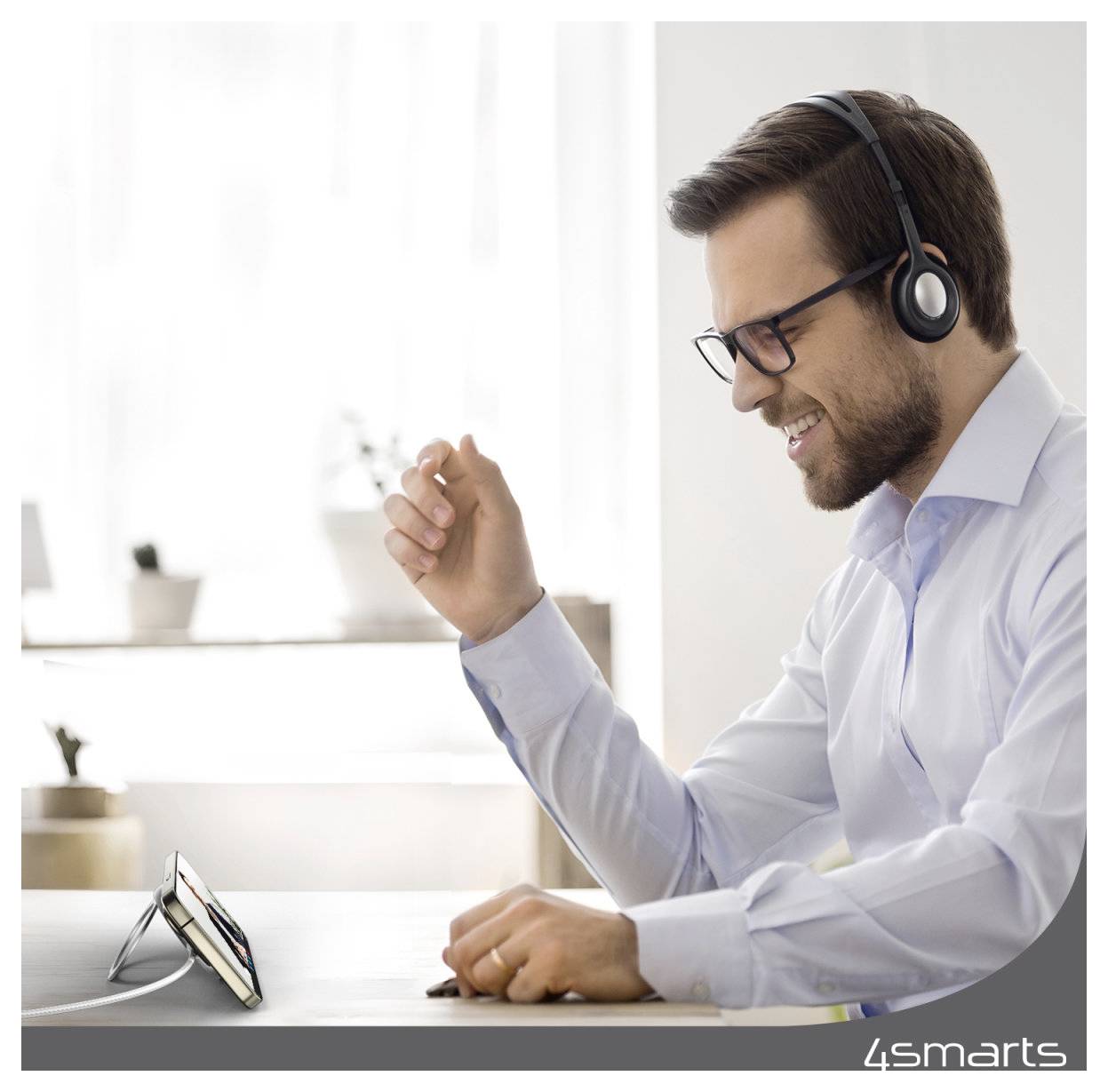 Lächelnder Mann mit Brille und Headset hebt die Faust während eines Videoanrufs, sitzt an einem Schreibtisch mit einem Smartphone auf einem Ständer.