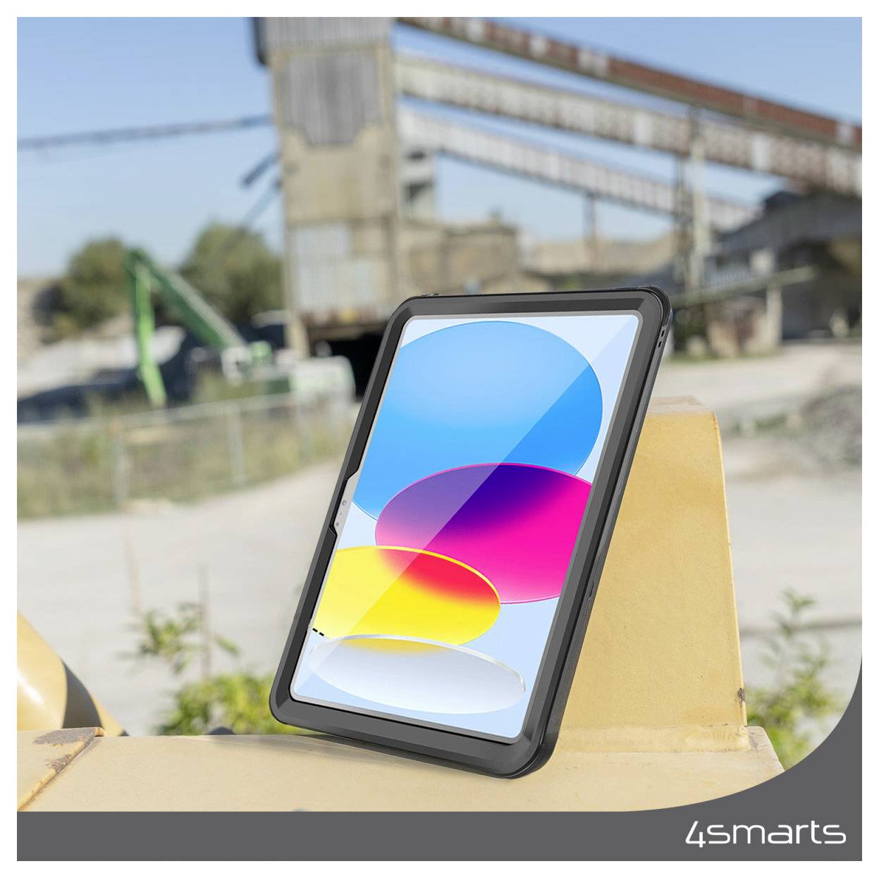 Tablet in einem robusten Gehäuse lehnt an einer gelben Barriere im Freien, mit einem farbenfrohen abstrakten Bildschirm; unscharfer Industriestandort im Hintergrund.