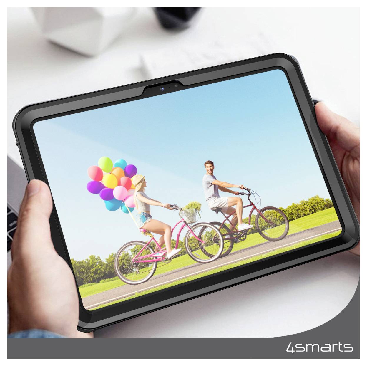 Hände, die ein Tablet halten, auf dem ein Paar zu sehen ist, das draußen Fahrrad fährt, wobei die Frau bunte Luftballons trägt.
