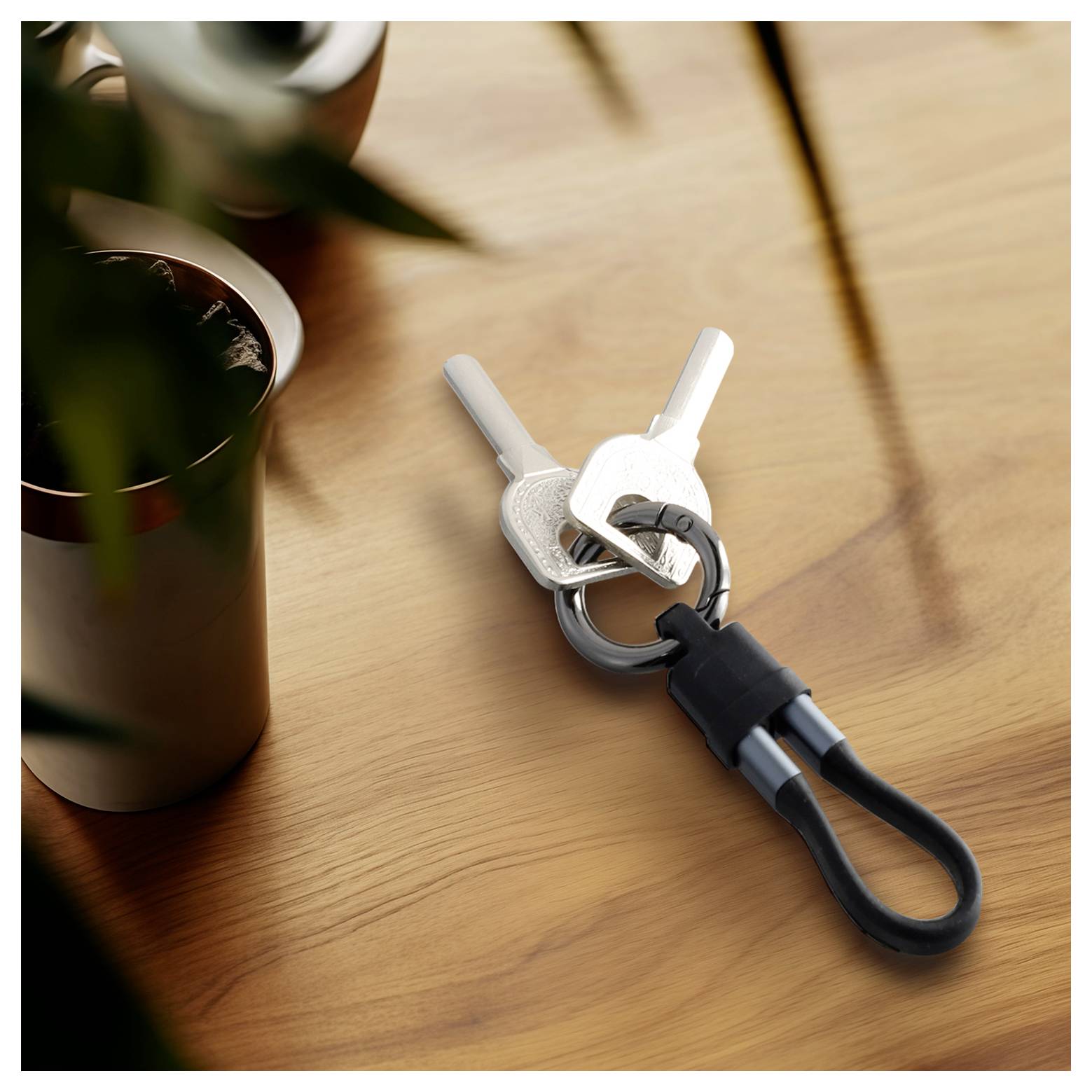 Zwei Schlüssel an einem Ring, befestigt an einem schwarzen Karabiner, liegen auf einem Holztisch neben einer Kaffeetasse.
