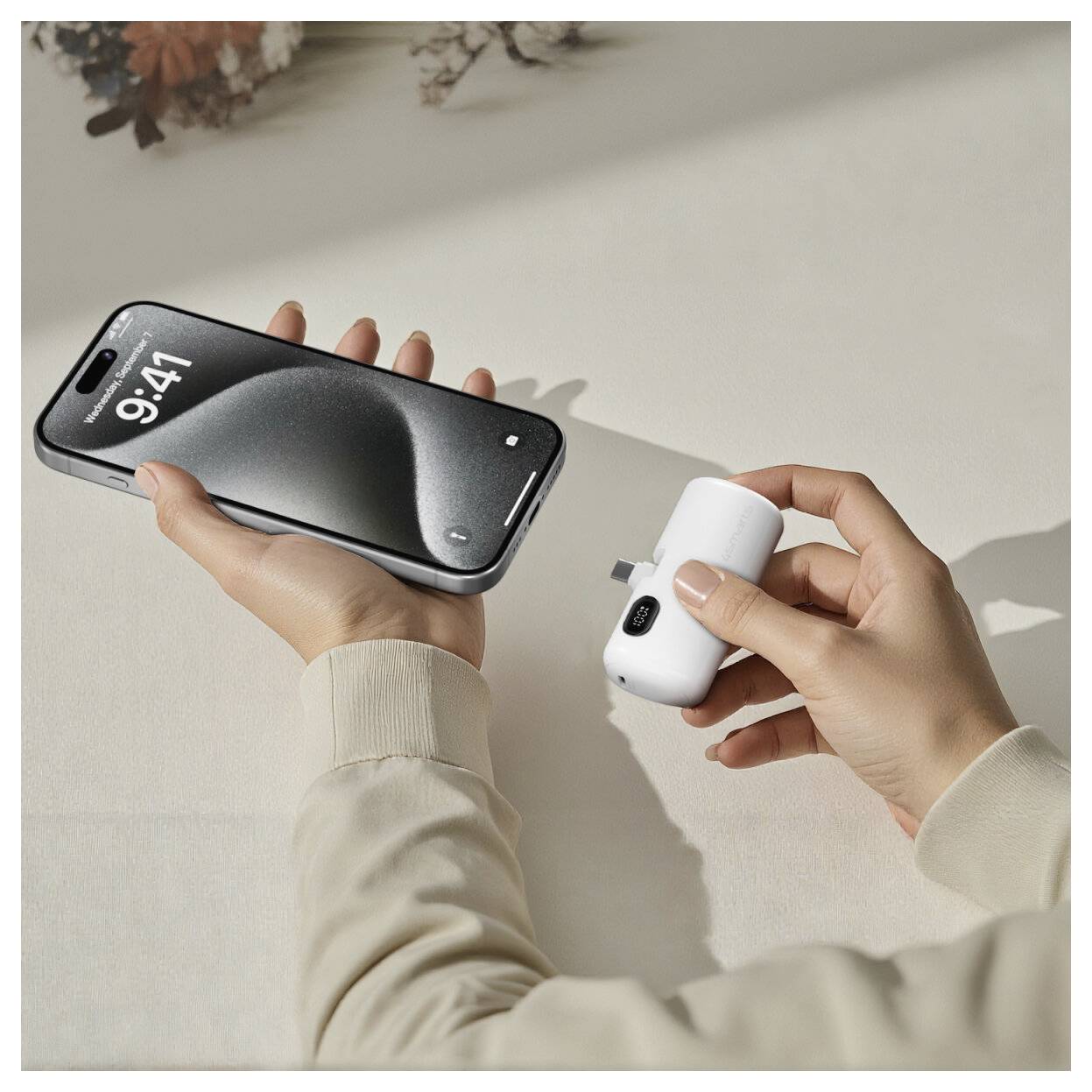 Hände halten ein Smartphone und einen kleinen weißen kabellosen Ladegerät auf einer hellen Oberfläche in der Hand, was auf mobiles Laden hindeutet.