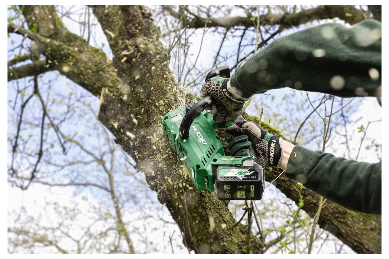 Handschuhgetragener Arbeiter, der eine grüne akkubetriebene Kettensäge verwendet, um einen Baumast im Freien zu schneiden.